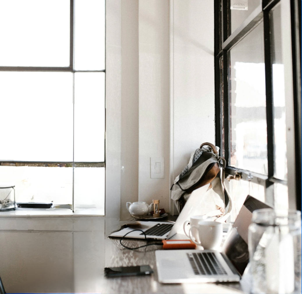 A workspace with two laptops, a mug, a teapot, and various items on a windowsill, with natural light coming through large windows.