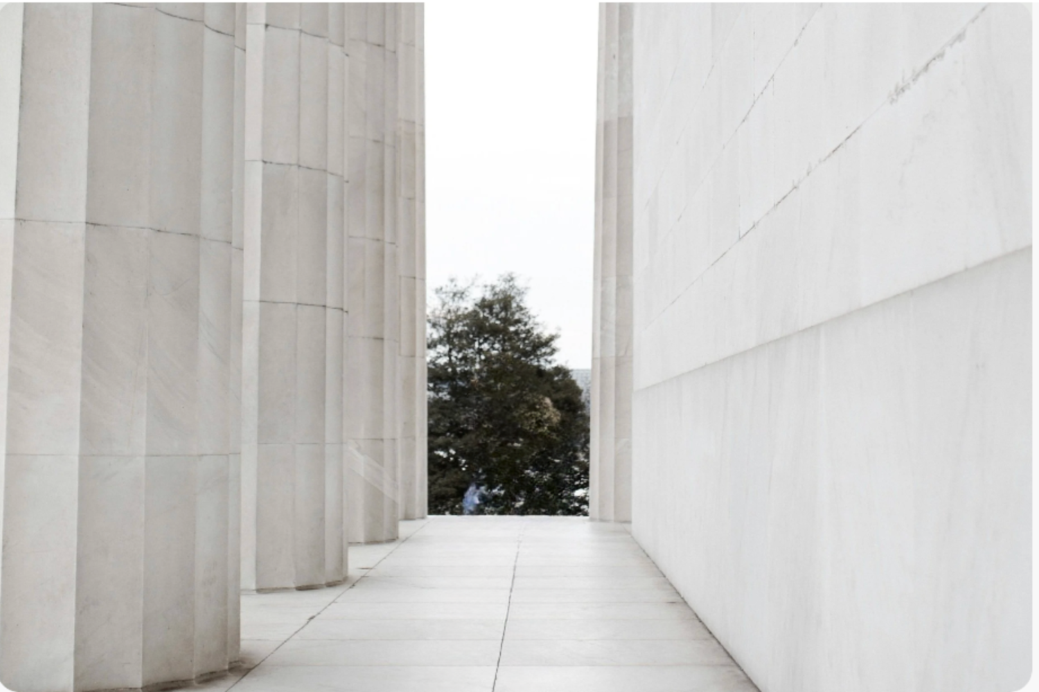 View through a narrow corridor between white marble columns and walls, with a tree visible in the background.
