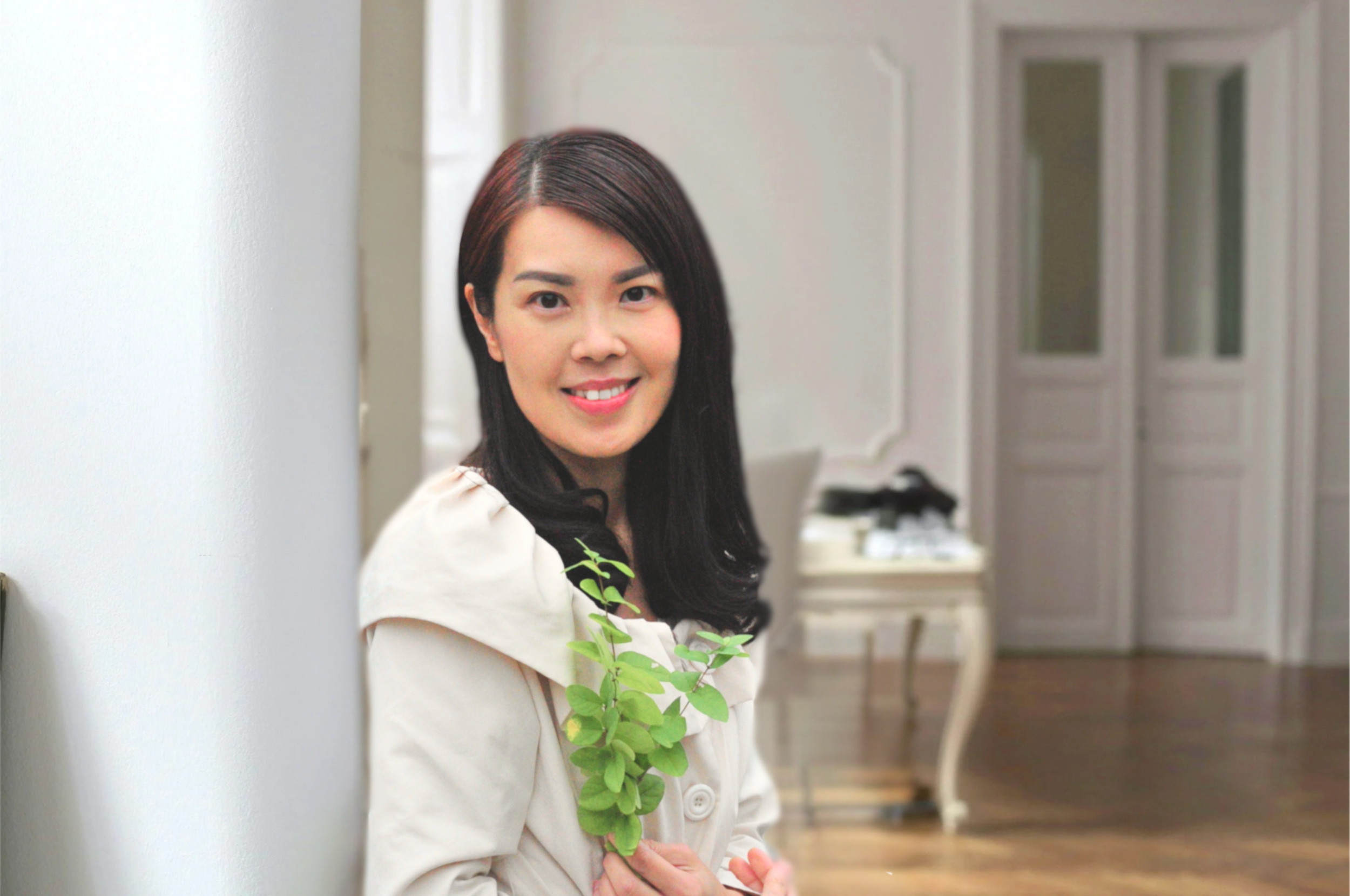 A woman with dark hair and a white blazer holding a green leafy branch while standing indoors in front of a white wall, with a blurred background of a room with white double doors.