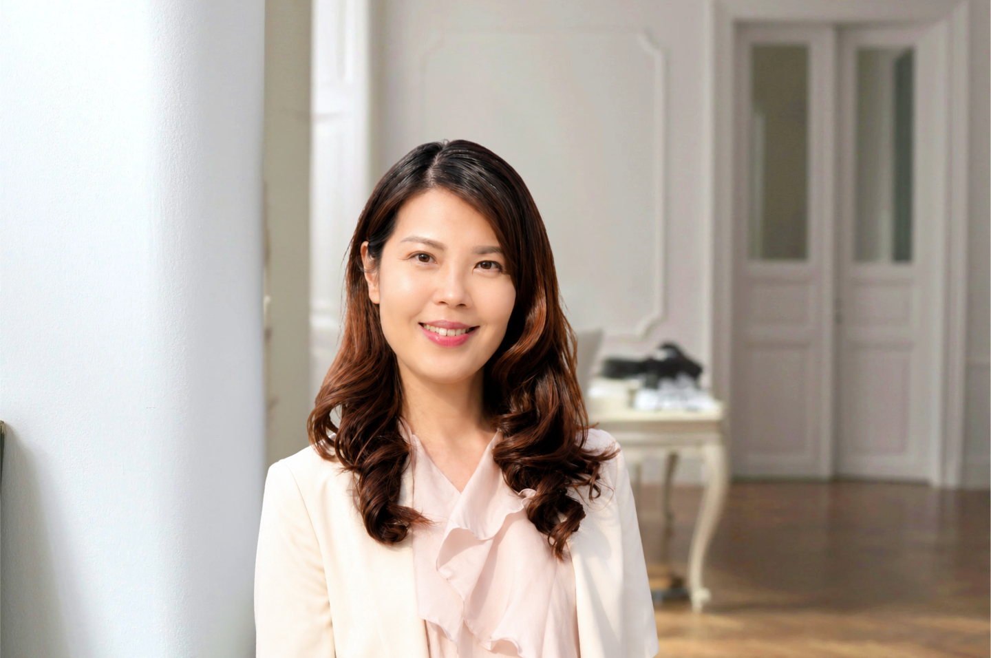A smiling woman with shoulder-length wavy dark hair, wearing light-colored clothing, standing in a bright, elegant room with white walls, ornate door frames, and a vintage-style table in the background.