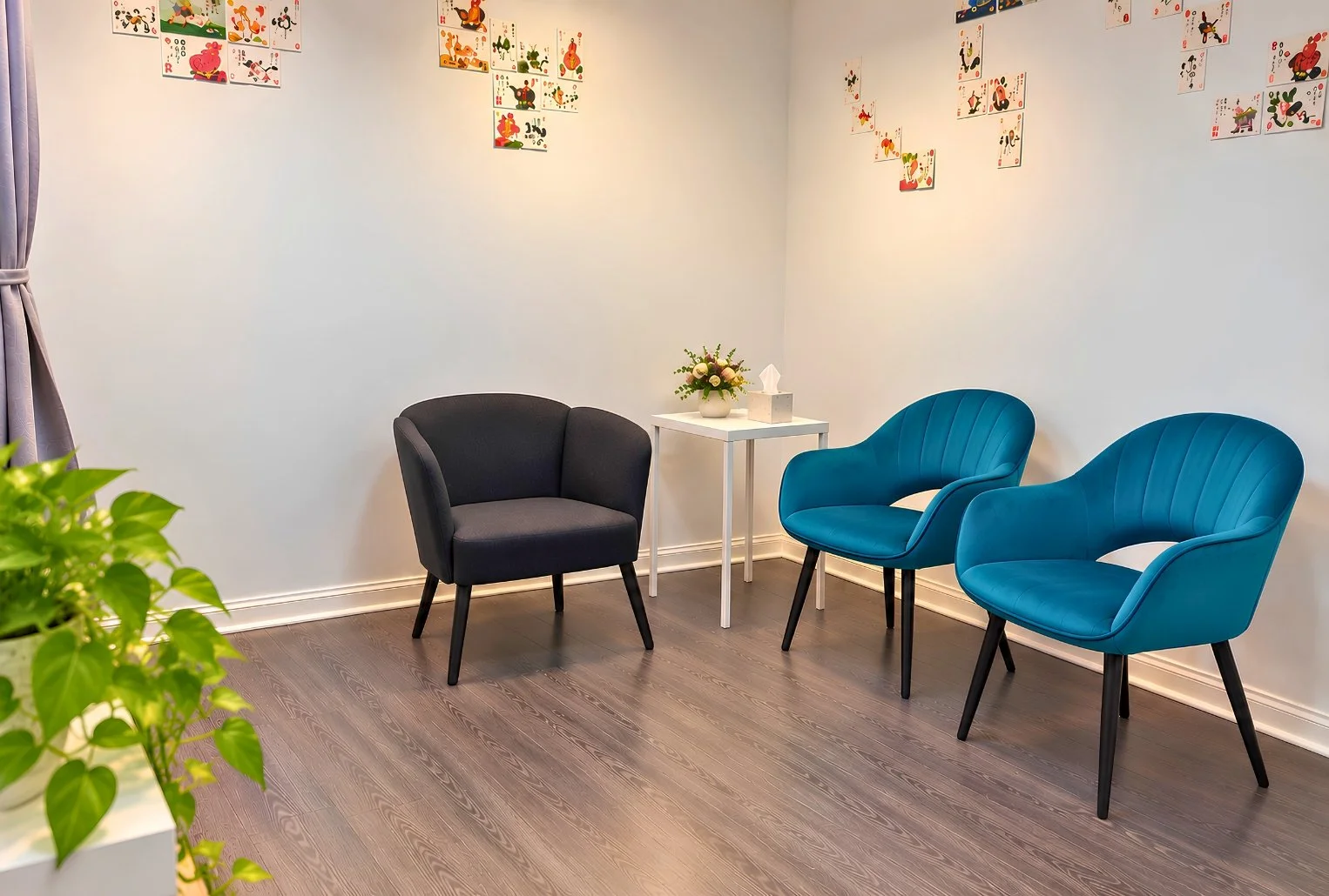 A waiting room with three chairs: one dark gray and two teal, arranged around a small white table with a flower arrangement and tissue box. Wall decor features colorful, playful artwork.