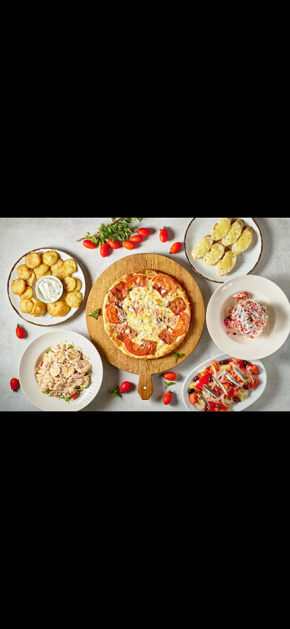 An assortment of food including pizza on a wooden paddle, potato salad, anchovy salad, cherry tomato salad, potato wedges, and a dessert with strawberries, presented on white and decorative plates and bowls on a light gray surface with scattered cherry tomatoes and sprigs of herbs.