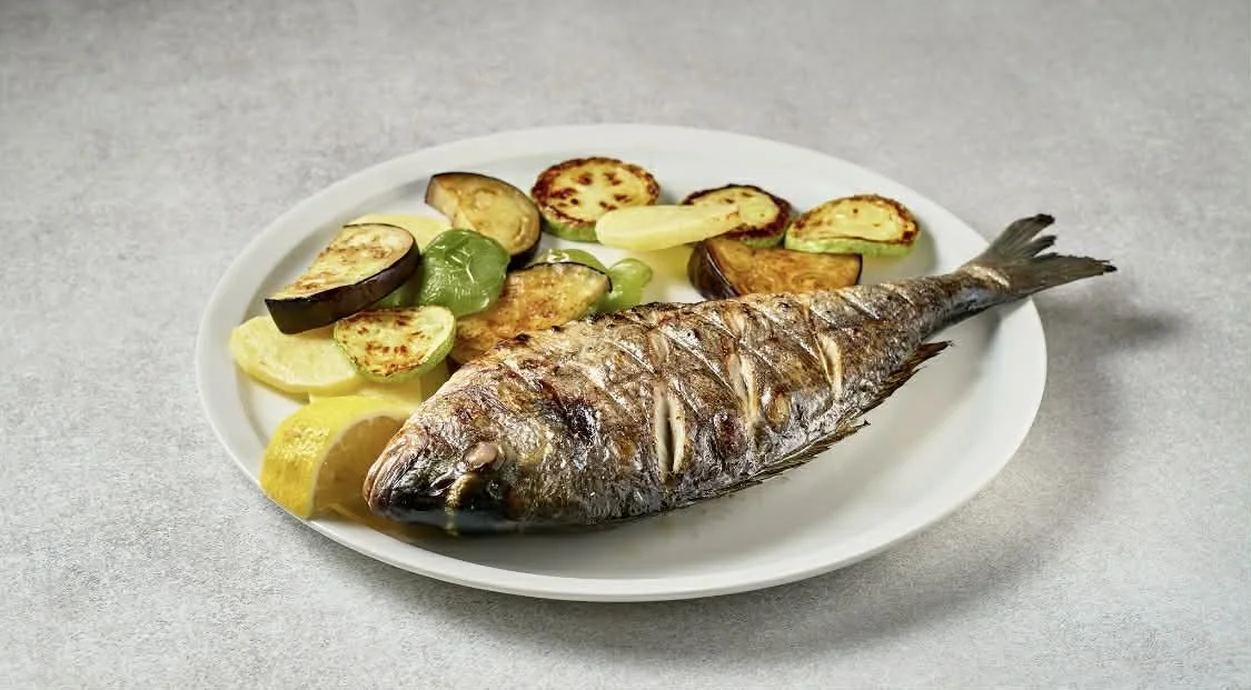 A whole cooked fish served on a white plate with roasted vegetables and lemon wedges.