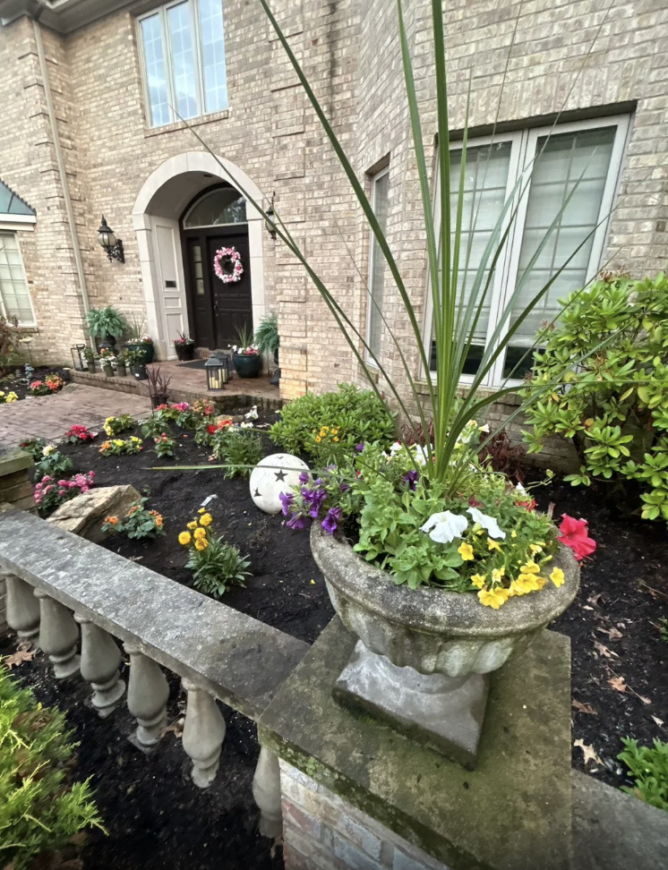 Front yard garden with potted plants, colorful flowers, and a brick house with a wreath on the door.