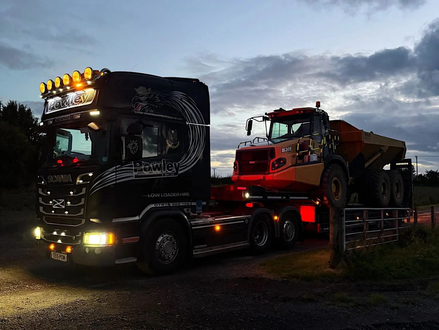 A large black and yellow dump truck loaded on a flatbed trailer, parked outdoors during dusk or dawn. The truck is marked with 'Powley' and has multiple lights on the front and top. The background has a cloudy sky.