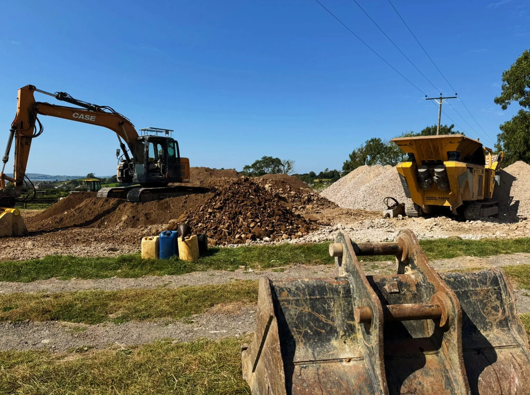 Construction site with an excavator and a bulldozer on dirt mounds, with grass and gravel foreground, and a utility pole with power lines in the background.