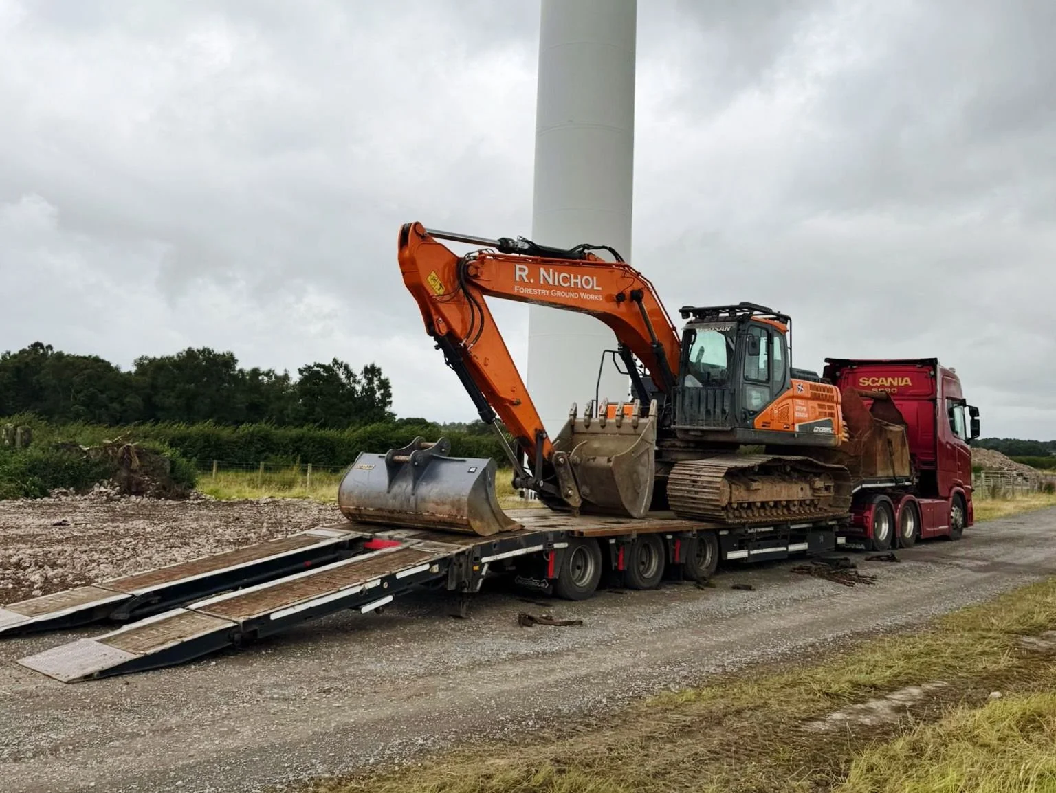 A construction excavator loaded onto a flatbed truck, situated outdoors on a gravel road, with a wind turbine and cloudy sky in the background.