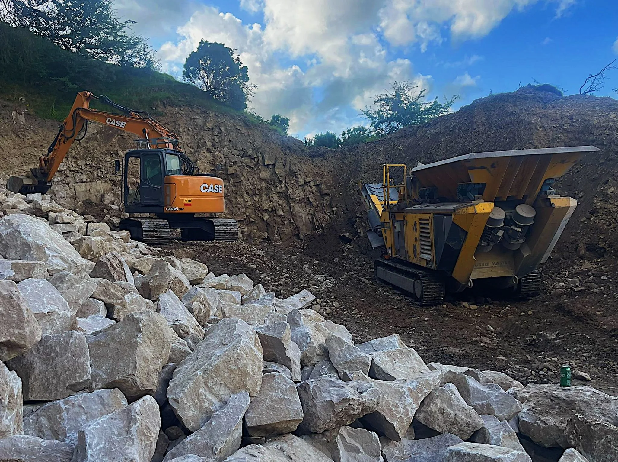 View of construction site with an excavator and a rock crusher working on a hillside, with rocks and dirt in the foreground and a partly cloudy sky above.