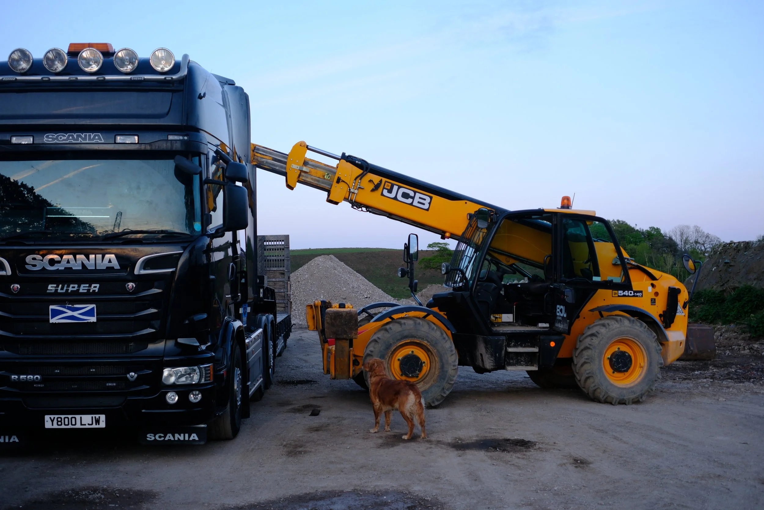 A black Scania truck with a Scottish flag on the front, a yellow JCB backhoe loader, a brown dog standing near the truck, and a small dog lying on the ground in front of the backhoe, all set outdoors with a dirt ground and a landscape background.