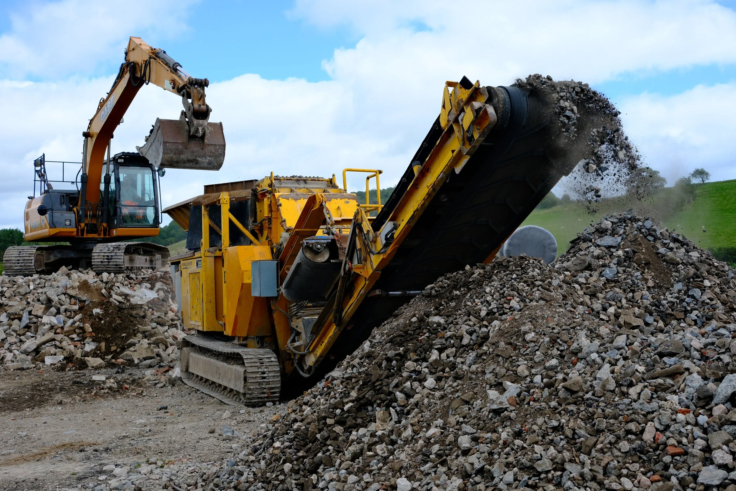 A construction excavator is dumping rocks onto a pile in an outdoor setting with green hills and a cloudy sky.