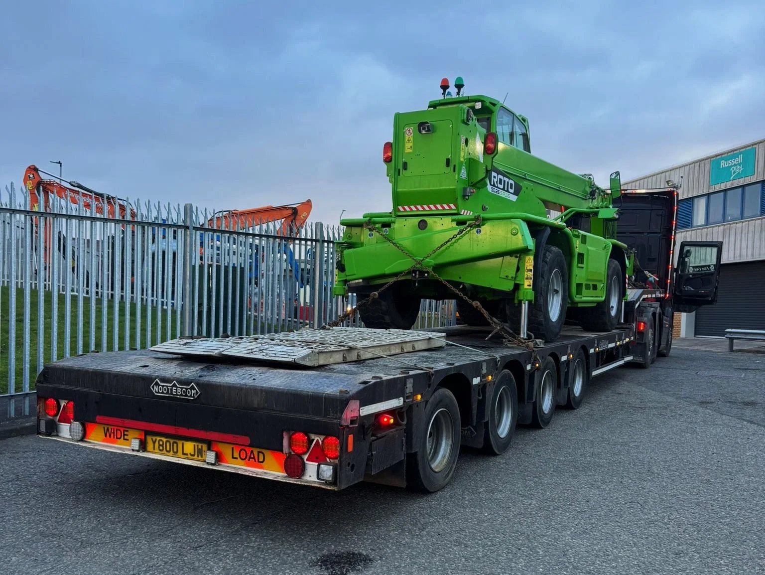 A green self-propelled construction vehicle loaded on a black flatbed truck parked outside a building with a sign that reads Russell Play. The vehicle has chains securing it to the truck and is surrounded by a metal fence, with orange excavators visi