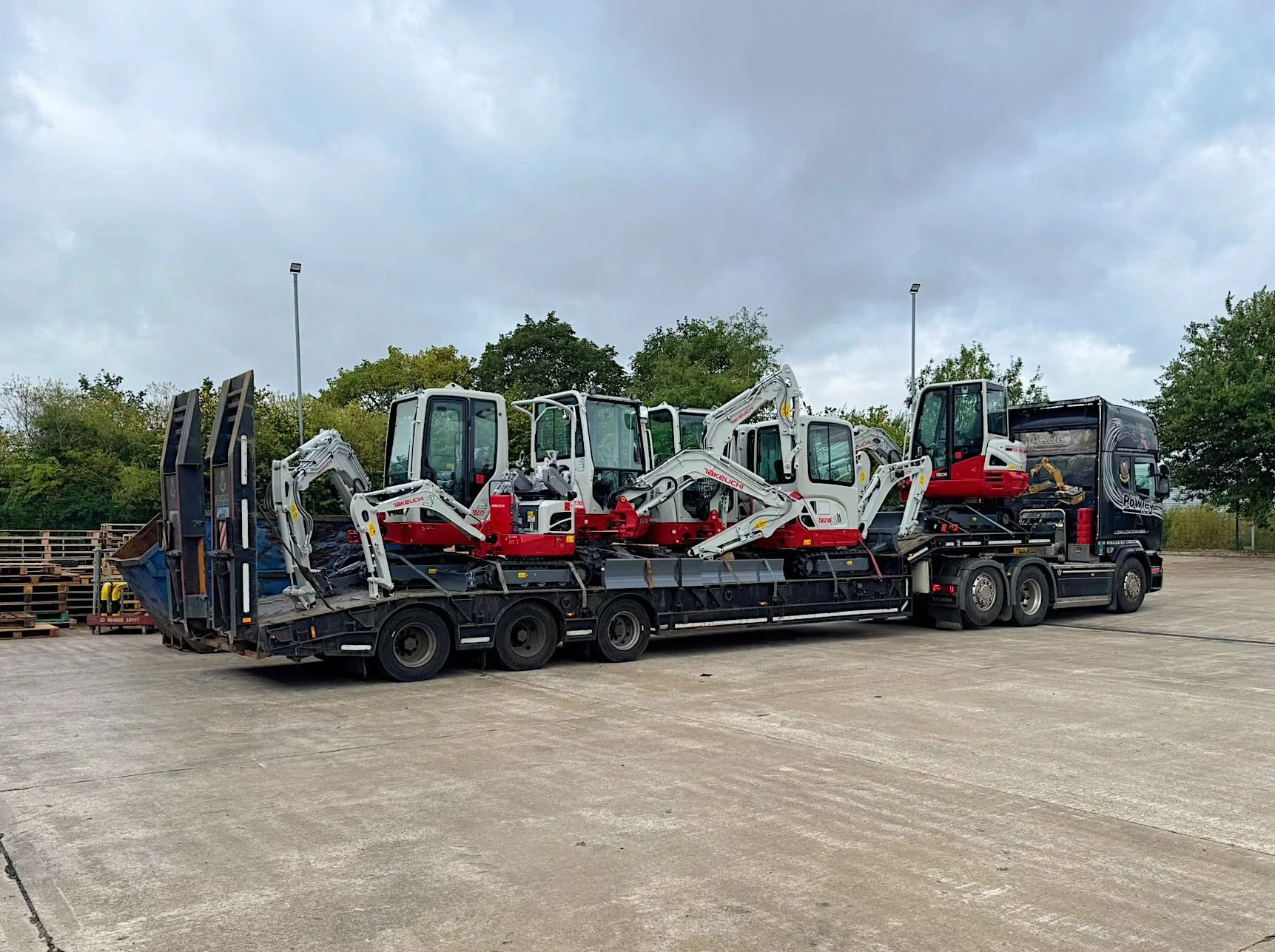 Flatbed truck carrying four mini excavators on an open lot with trees and cloudy sky in the background.