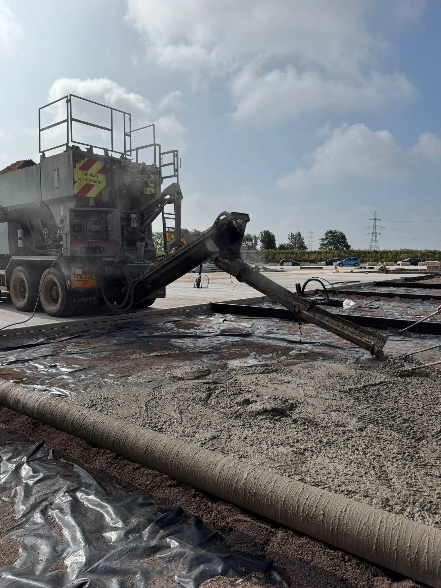 A large construction vehicle laying wet concrete on a road, with several cars in the background and power lines in the sky.