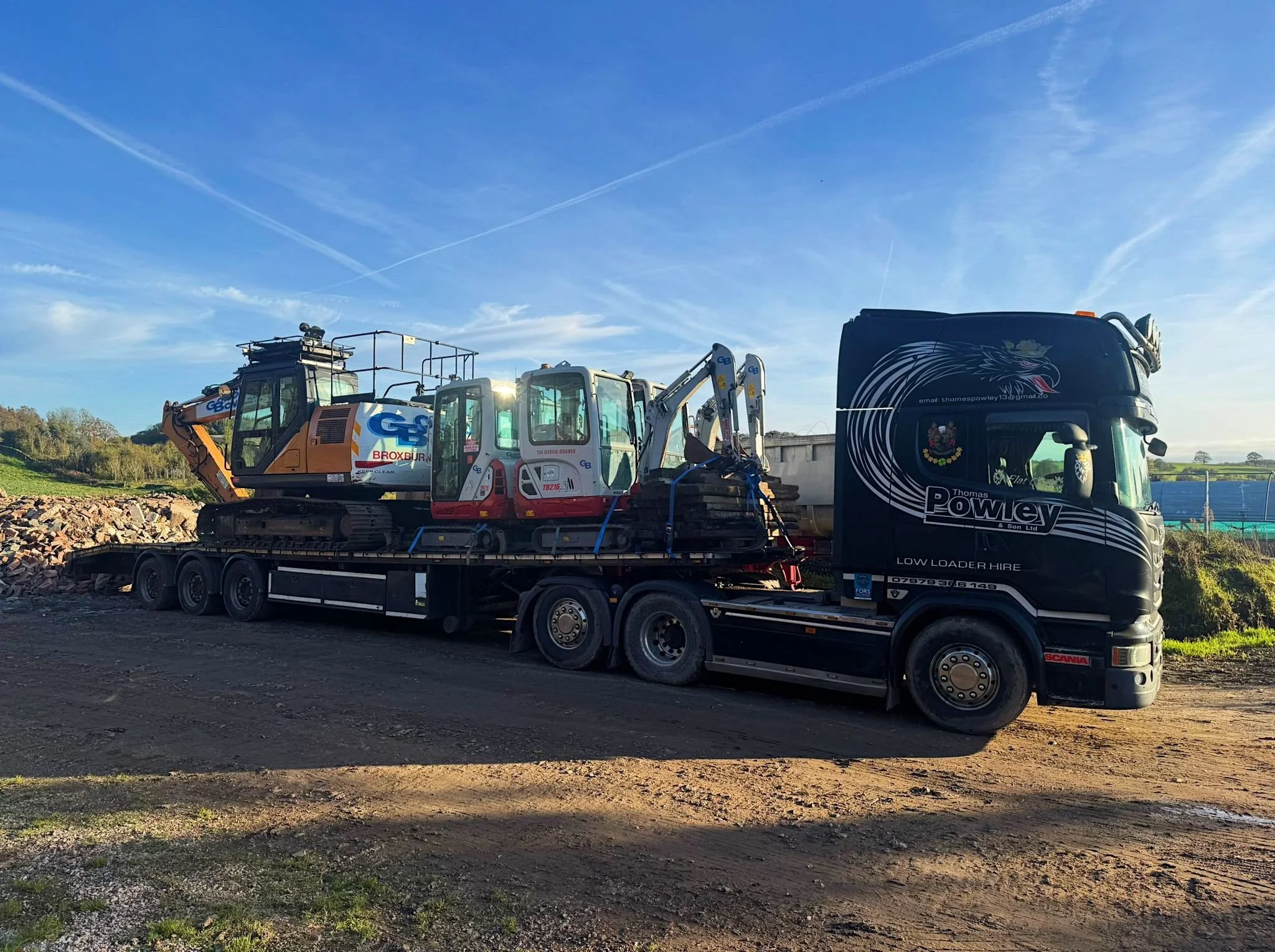 A large black truck from Thomas Powley & Son Ltd. is parked on a dirt area and carrying a construction excavator on its trailer. The excavator has a white body with red and blue markings and is labeled 'G & B Brookbourn.' The background features a bl