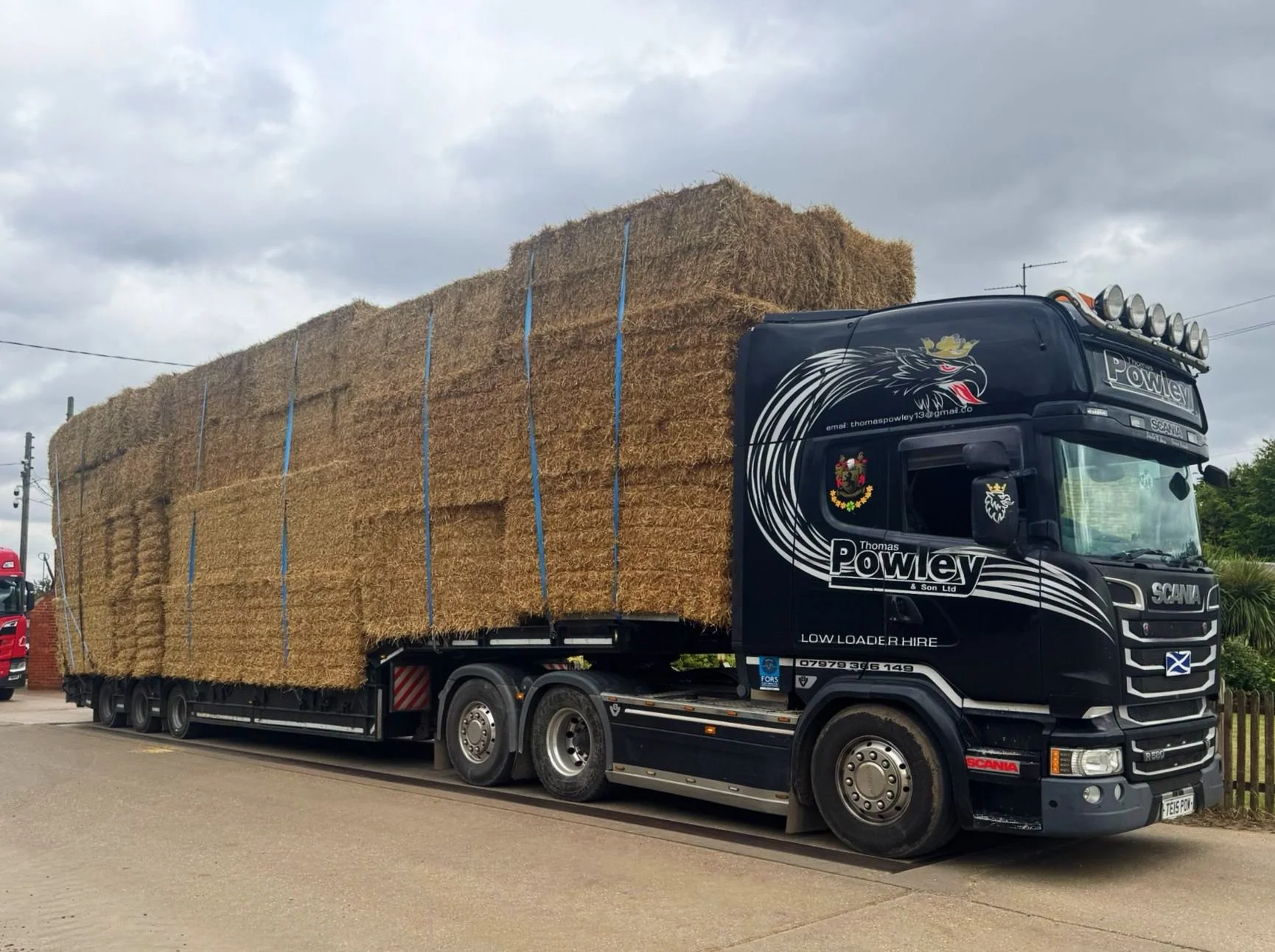 A large truck parked on the side of the road, loaded with stacks of hay bales, with a cloudy sky overhead.