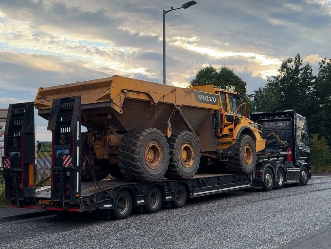 A large yellow Volvo construction vehicle, likely a dump truck or loader, on a flatbed trailer truck, being transported on a highway with trees and a cloudy sky in the background.