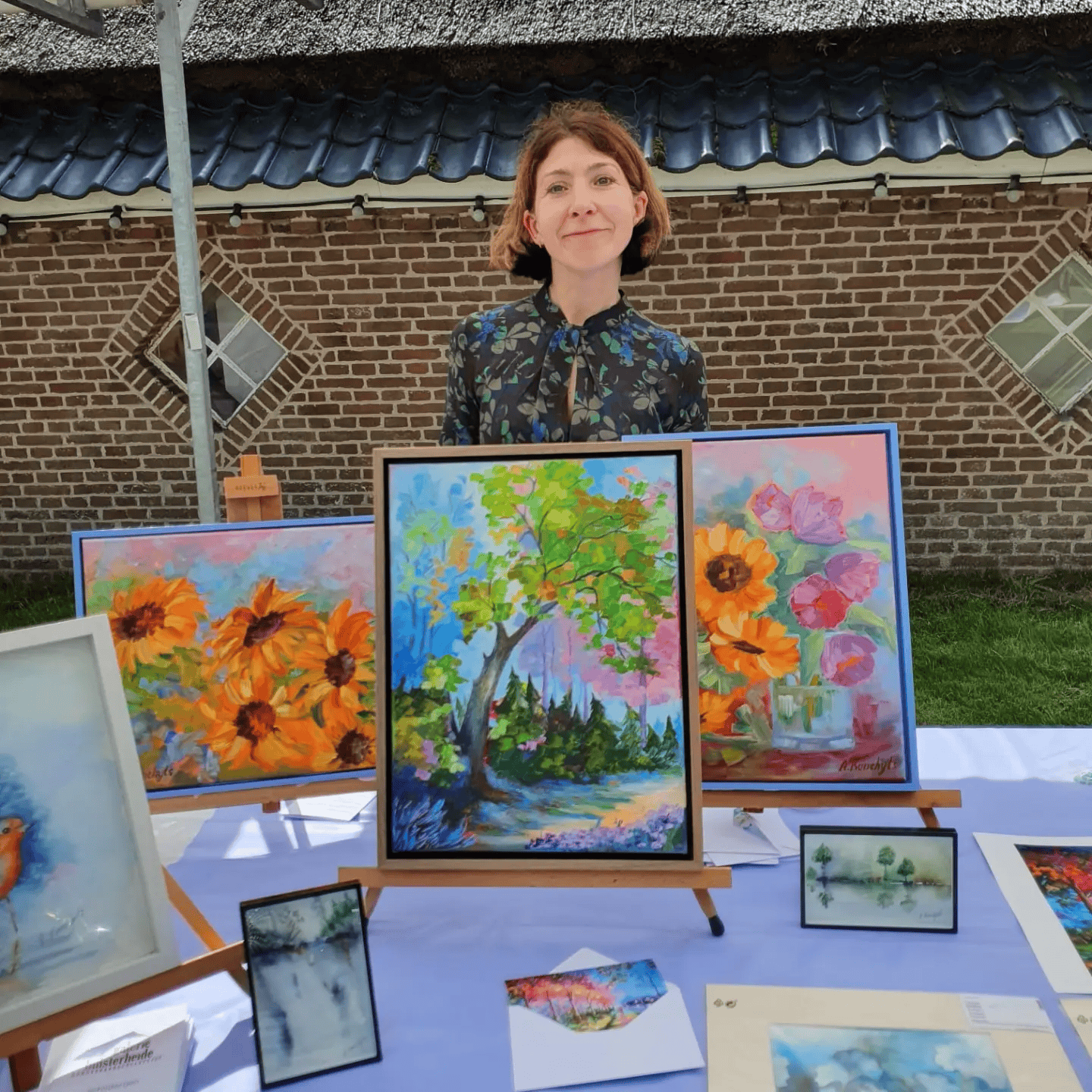 A woman standing behind a table displaying colorful landscape and floral paintings outdoors, with a brick building and two diamond-shaped windows in the background.