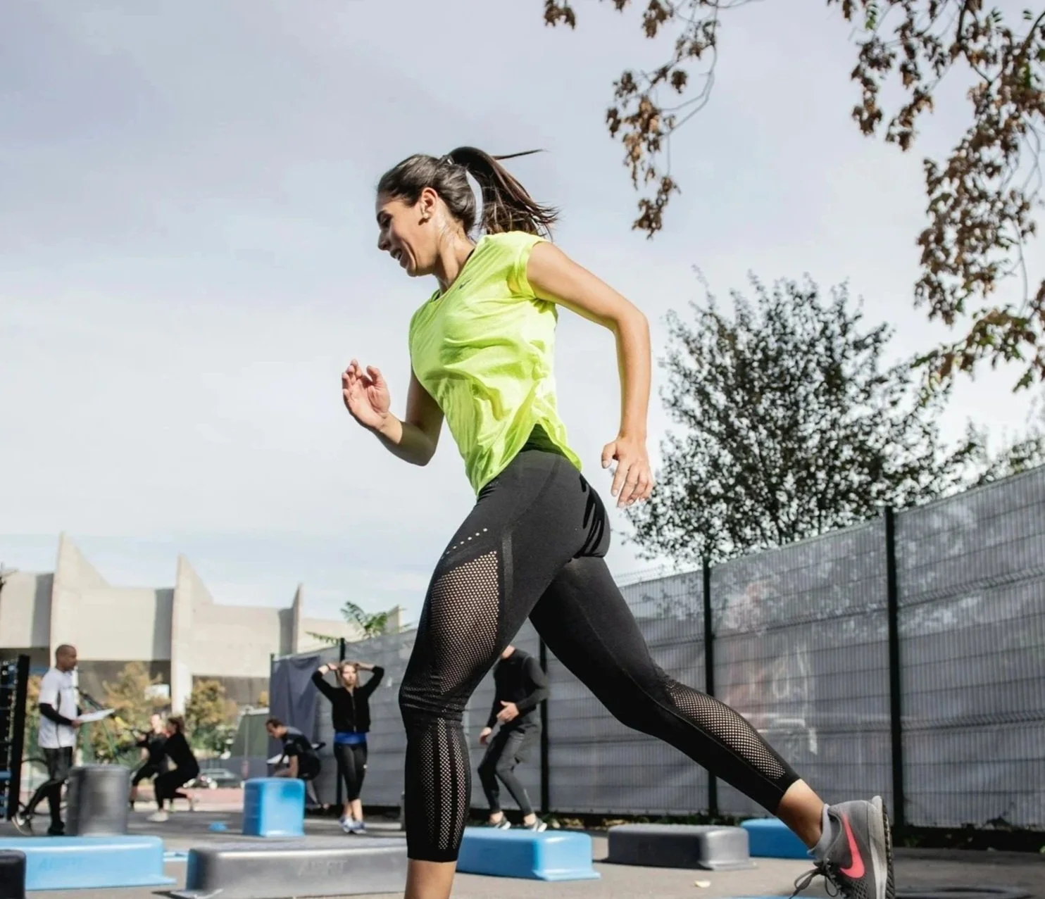 A woman running outdoors in athletic gear, wearing a yellow top and black leggings with mesh panels, on a cloudy day with trees and a fence in the background, and other people exercising nearby.