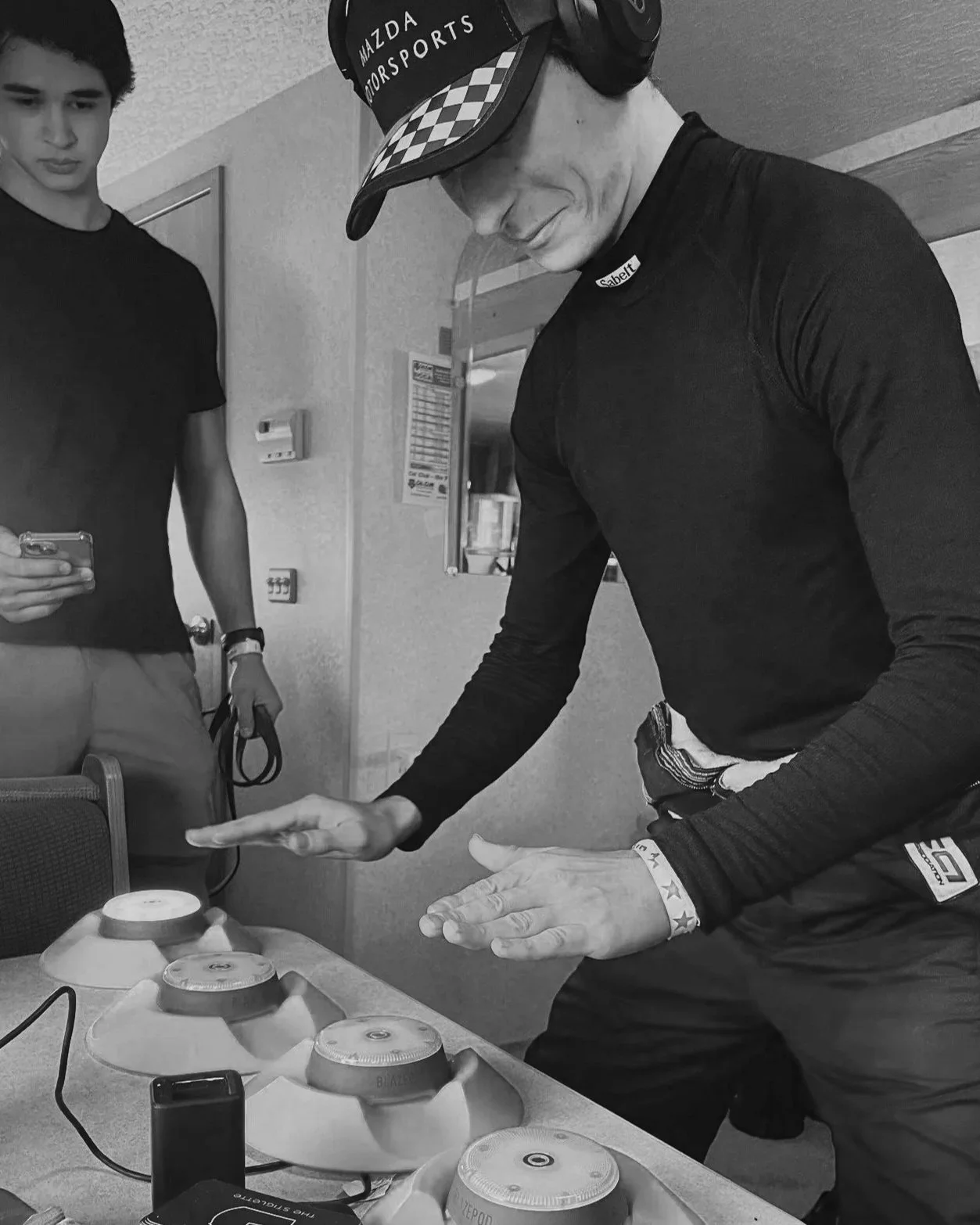 Person wearing a racing cap and headphones demonstrating a technique with a spinning top at a table, while another person observes and holds a phone in a room with various equipment.