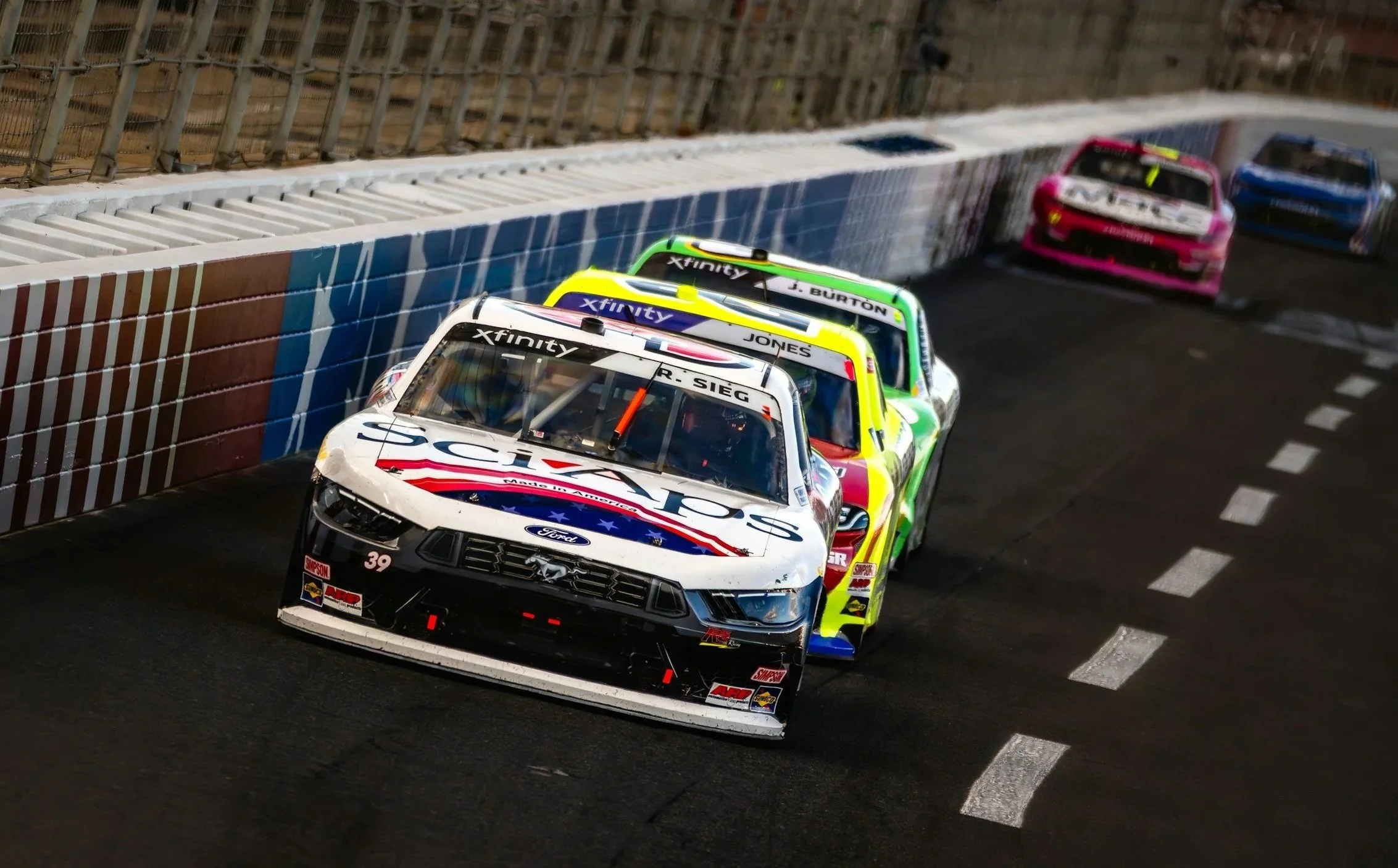 Race cars on a racing track, with the lead car displaying NASCAR sponsorship, followed by three other cars closely behind, and a brick wall with fencing along the side of the track.