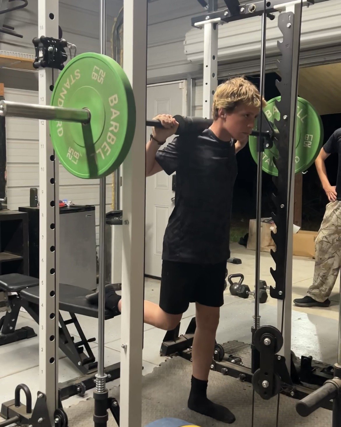 A young man in black athletic clothing performs a squat exercise with a barbell loaded with green weight plates in a garage gym.
