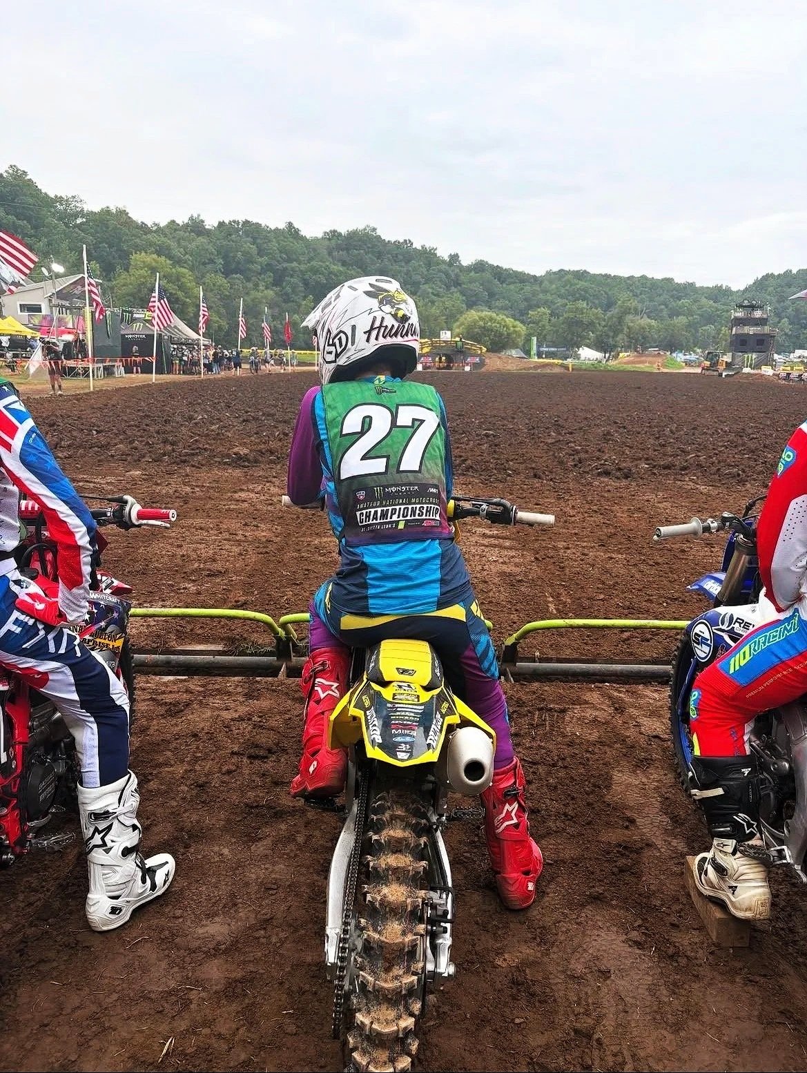 Motocross riders preparing on the starting grid at a dirt track, with one rider in the center wearing a green vest with the number 27, and others on either side in colorful gear. The track is muddy, with a crowd and American flags in the background.