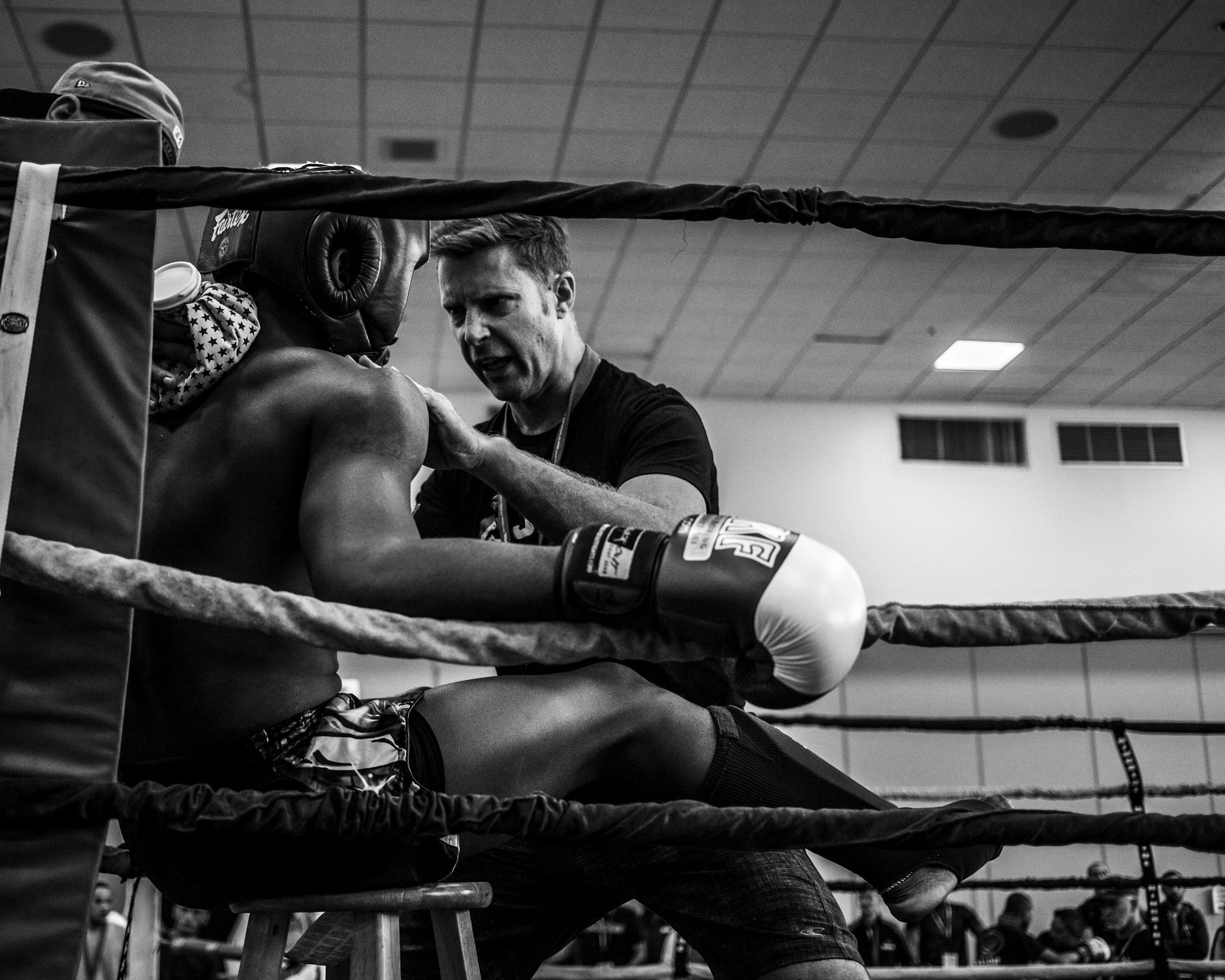 A boxing trainer leaning over a young boxer in a boxing gym, offering encouragement or instructions. The scene is in black and white with visible boxing ring ropes.