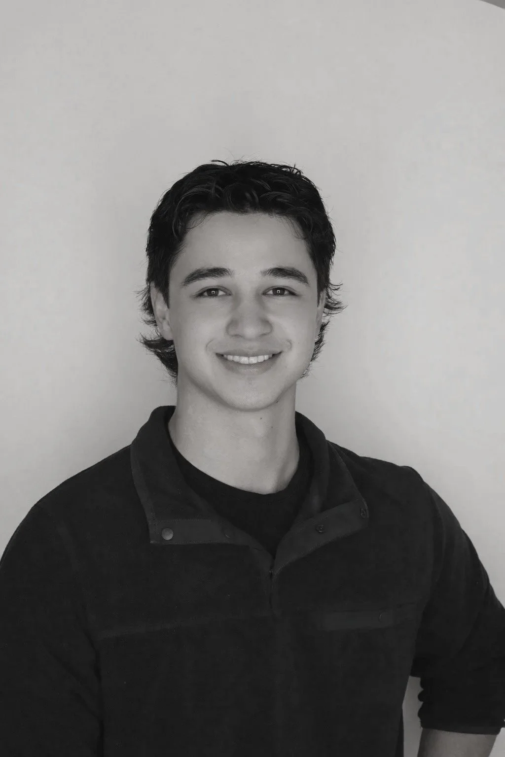 Black and white portrait of a young man with dark wavy hair, smiling, wearing a dark collared shirt against a plain light background.