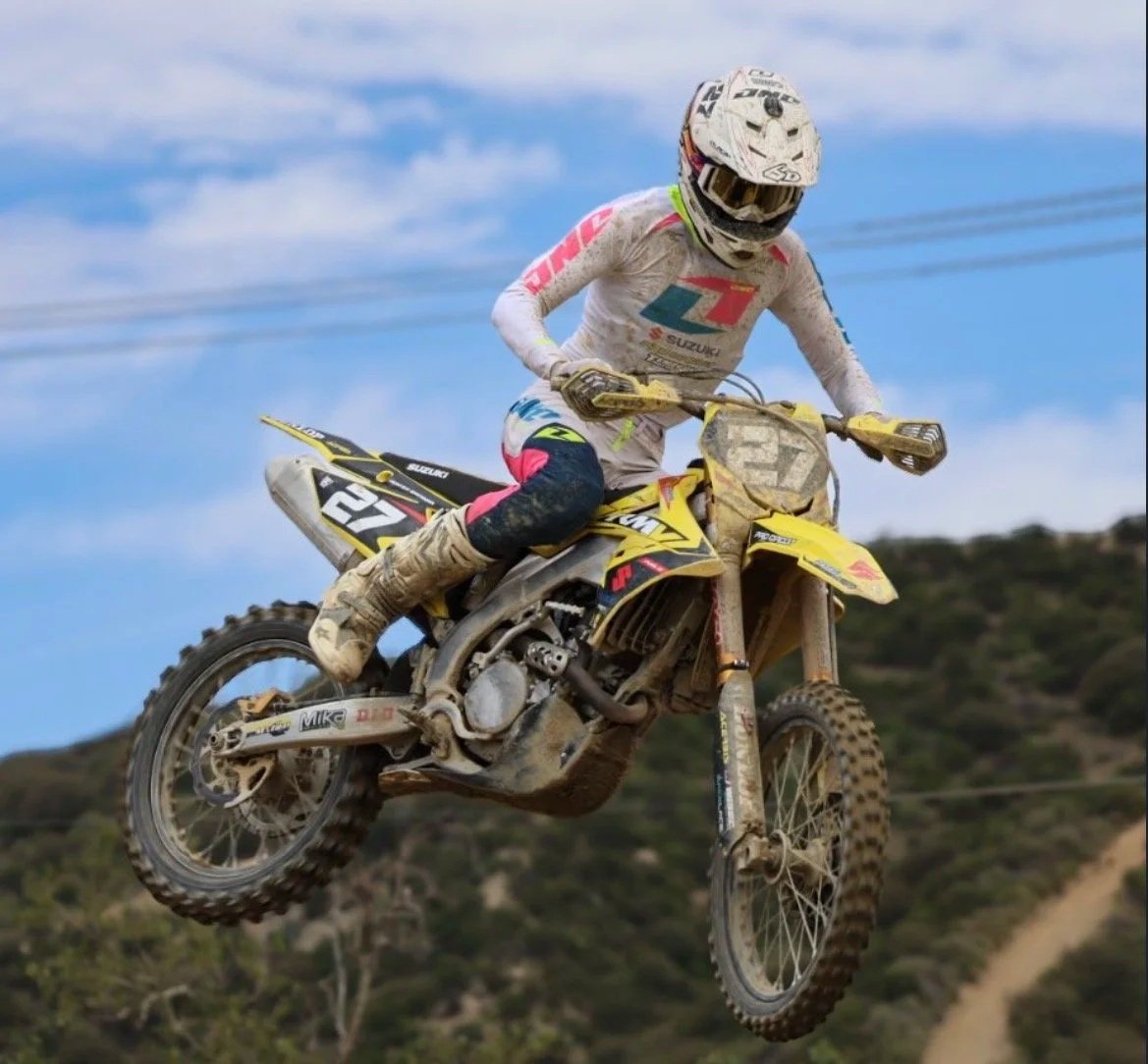 Motorcyclist jumping mid-air on a dirt bike, wearing a white helmet and racing gear, with a background of hills and blue sky.