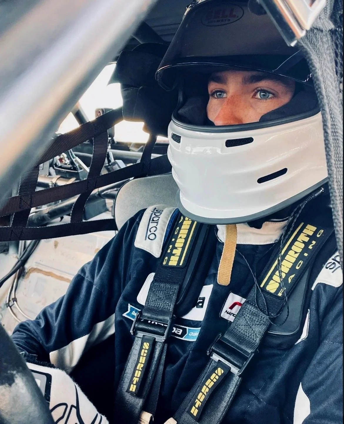 Close-up of a race car driver wearing a black racing suit with sponsor patches and a white helmet, seated inside a race car cockpit surrounded by safety harnesses.
