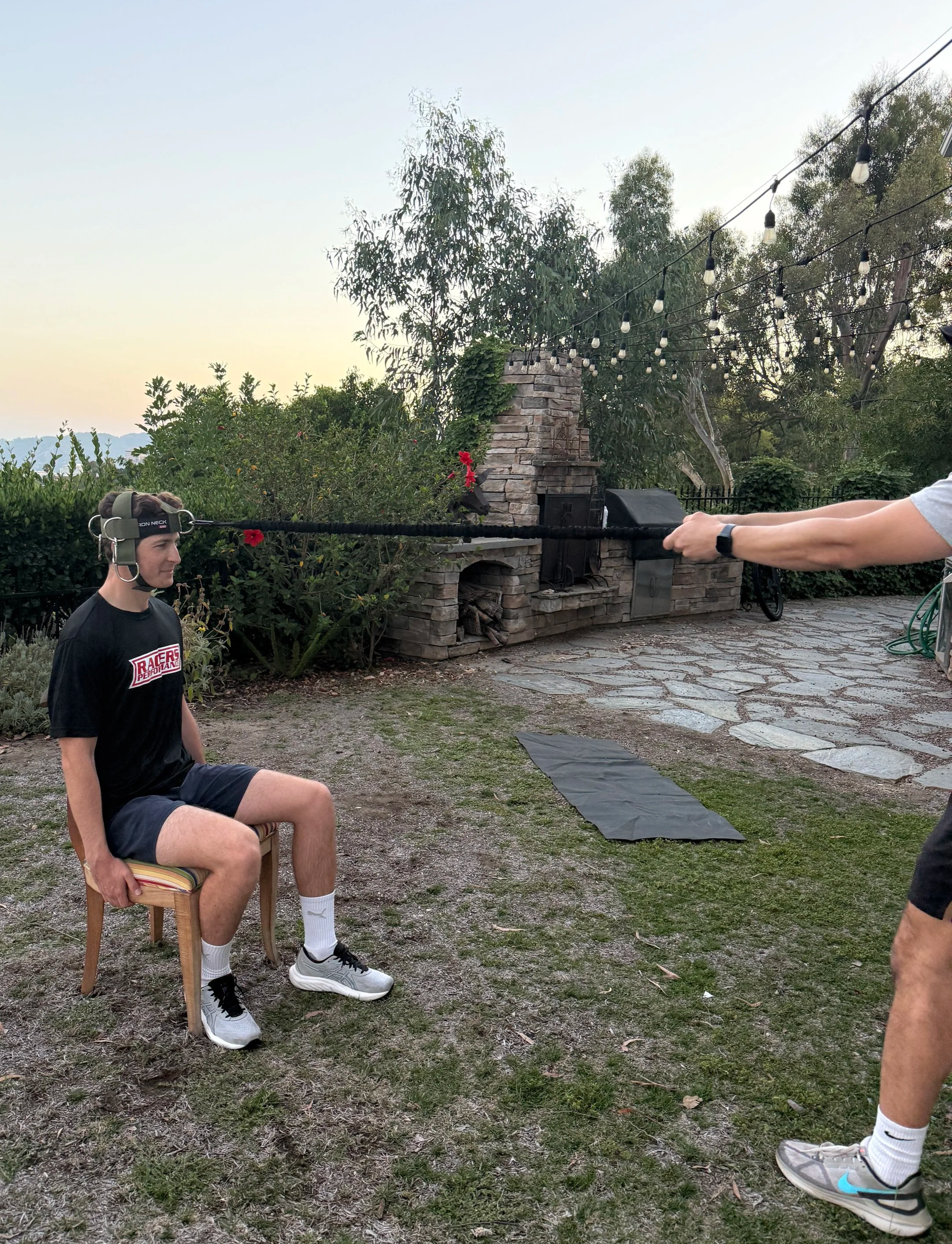 A person with a helmet and black athletic clothing is sitting on a wooden chair outdoors on a grassy area. Another person is standing in front of them, holding their hands with a resistance band, which is positioned around the seated person's shoulders. In the background, there is a stone fireplace, trees, string lights, and a paved area.