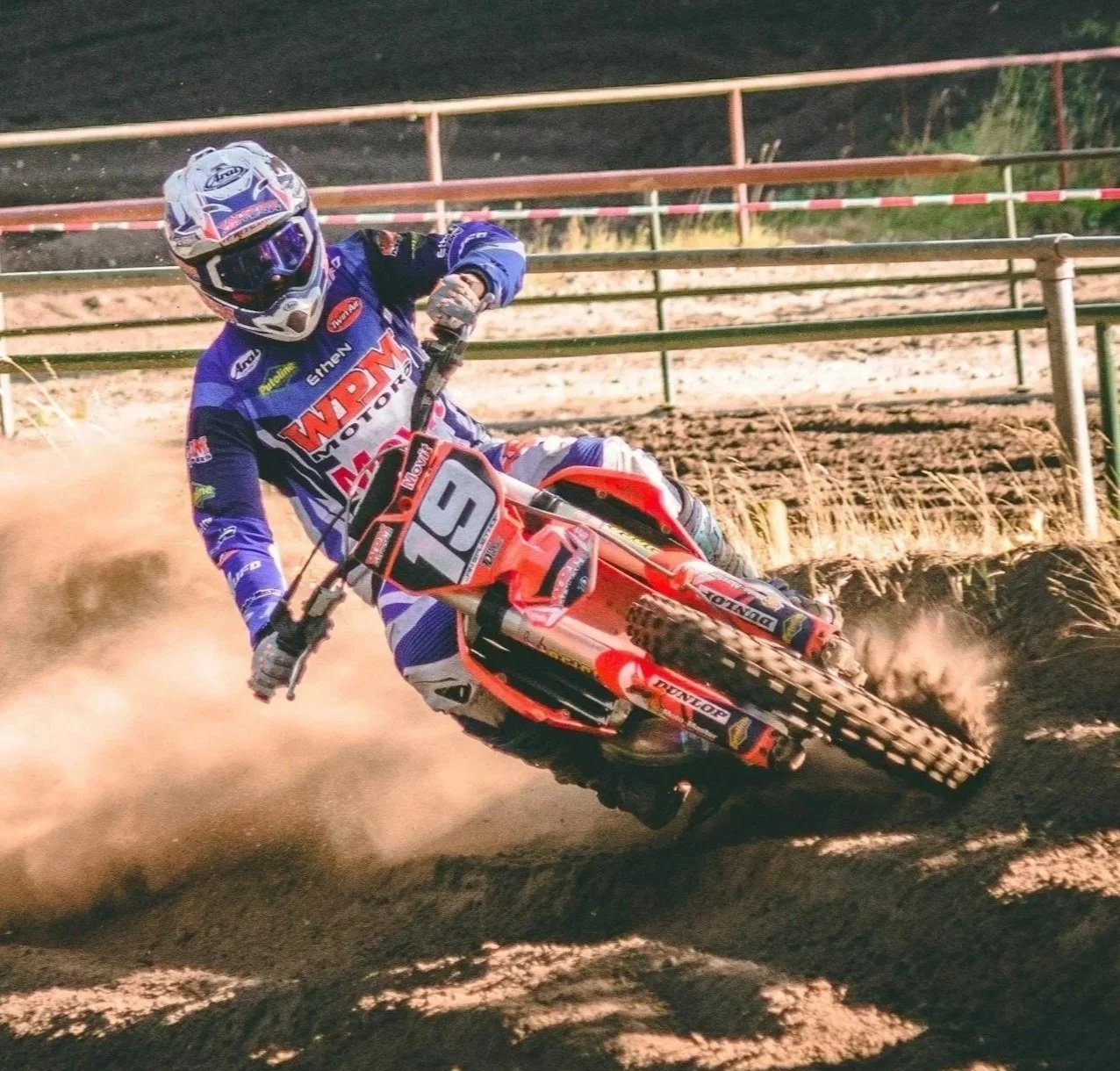 Motorcycle racer in blue racing gear and helmet leaning into a turn on a dirt track, kicking up dust with barriers in the background.