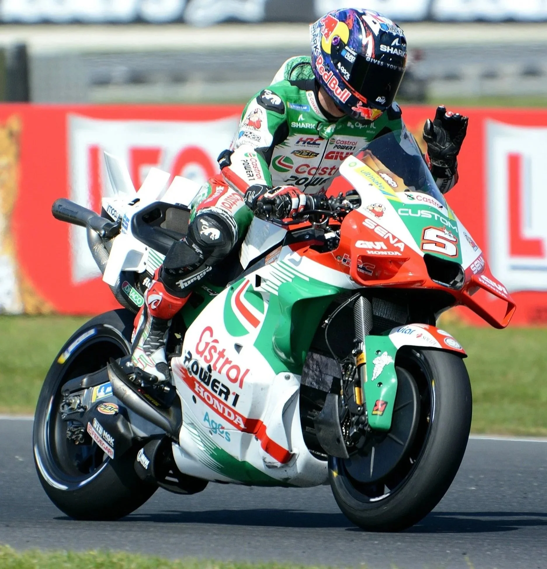 A motorcyclist wearing a colorful racing suit and helmet is riding a race bike on a paved track. The bike is decorated with sponsor logos and a green, white, and red color scheme. The rider appears to be celebrating or signaling with a raised hand.