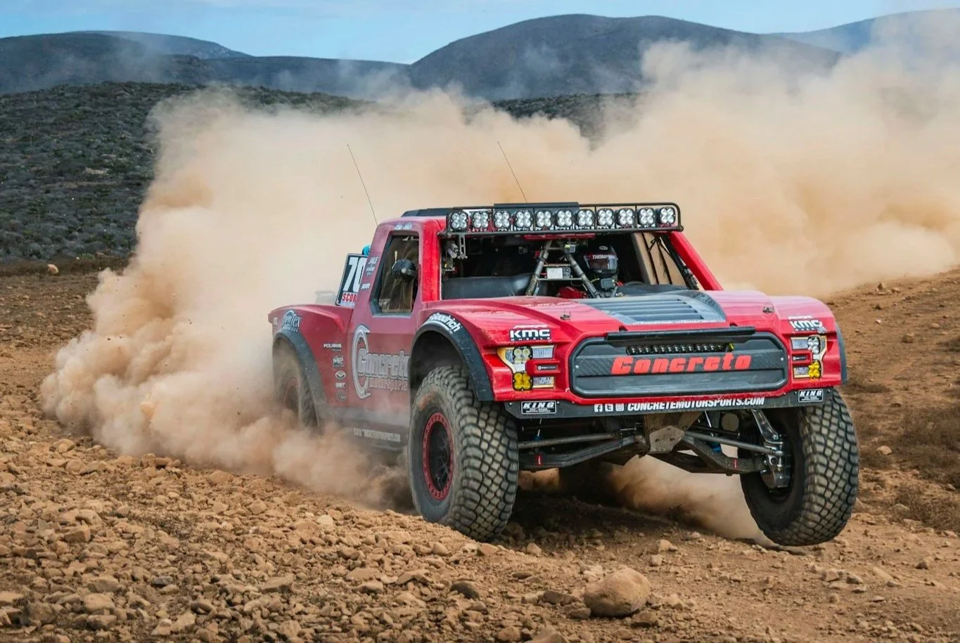 Red off-road racing truck driving on dirt terrain, kicking up dust, with mountains in the background.