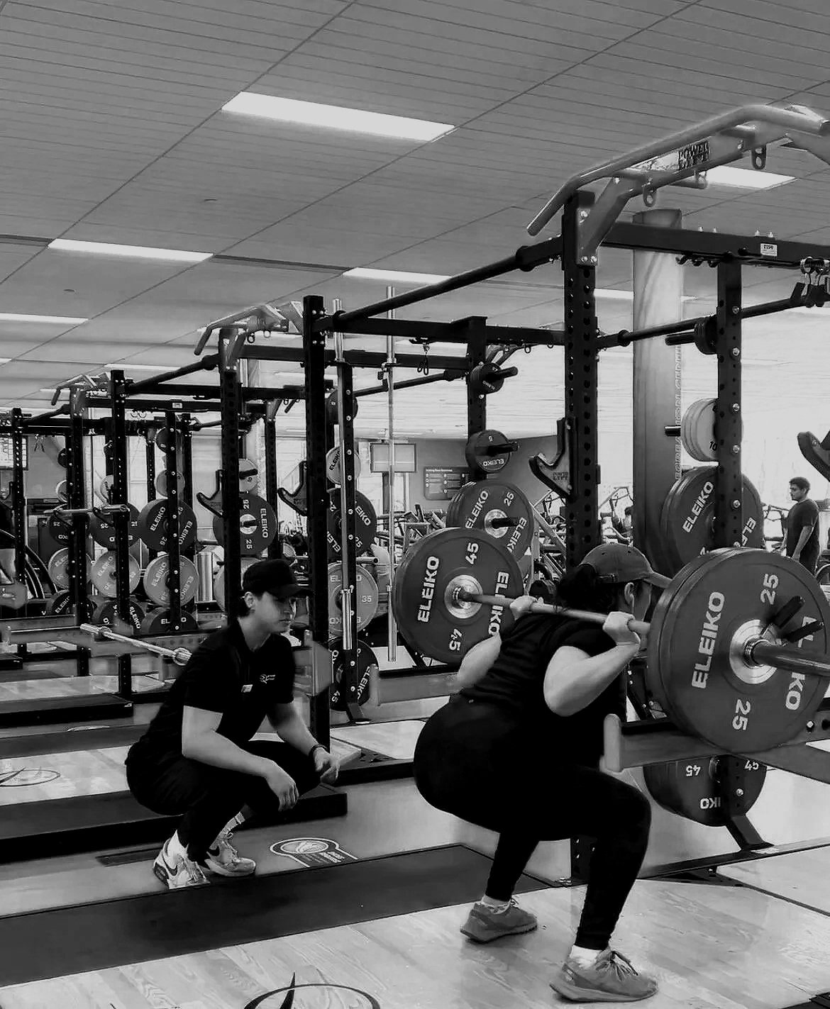 A person lifting weights in a gym, with a trainer kneeling nearby, surrounded by gym equipment including weight plates and racks.