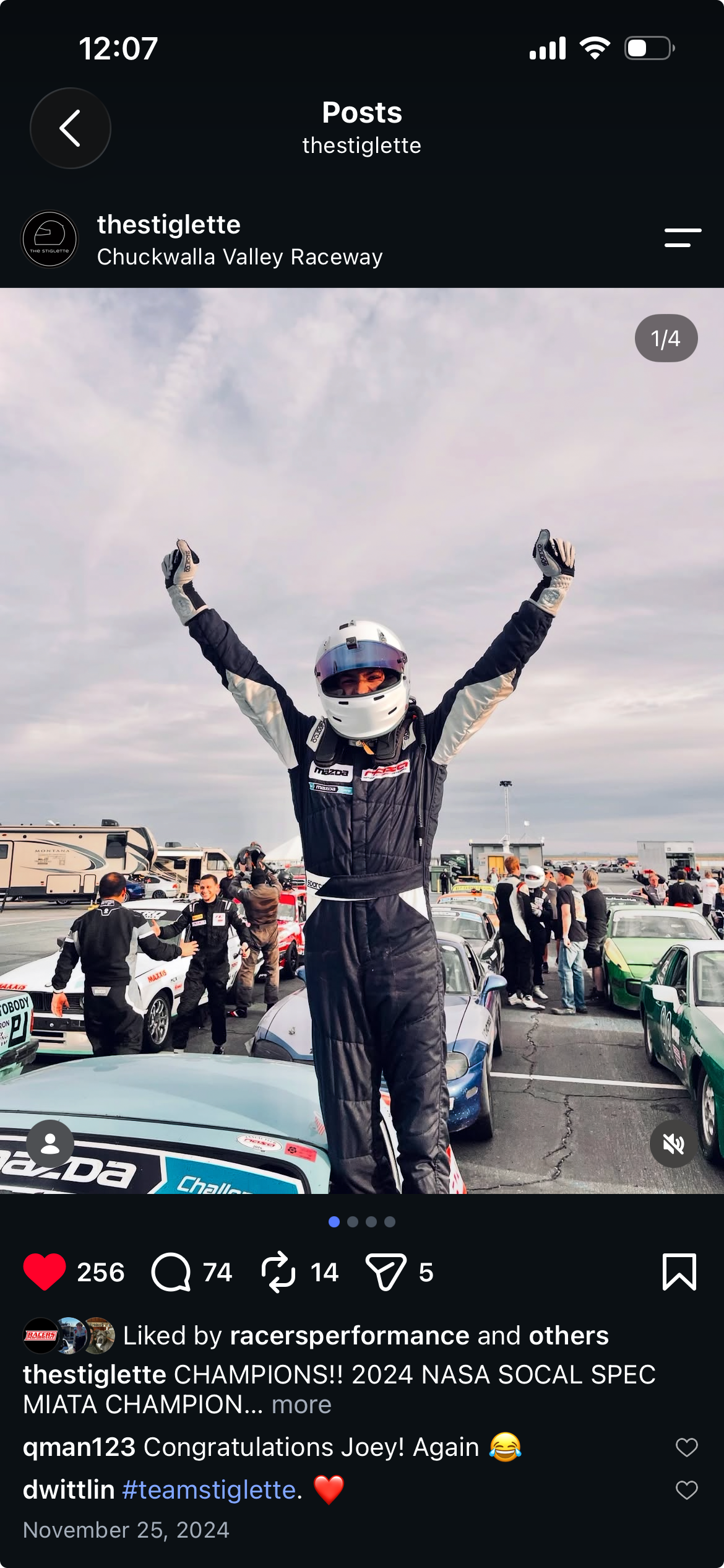 Race car driver in racing suit and helmet celebrating with arms raised at the track, surrounded by race cars and team members at Chuckwalla Valley Raceway.