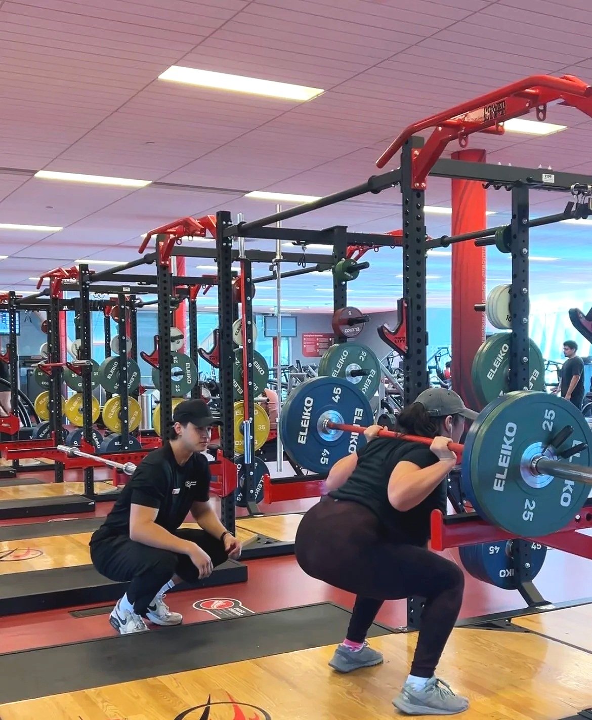 A woman performing a squat exercise with a barbell loaded with weight plates in a gym, with a trainer spotting her. The gym has multiple weightlifting stations, mirrors, and people in the background.