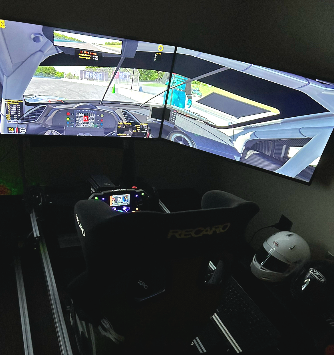 Sim racing setup with a large curved monitor displaying a race car cockpit view at a racing circuit, a racing wheel controller, a racing seat branded RECARO, a white racing helmet, and additional racing equipment on the desk.