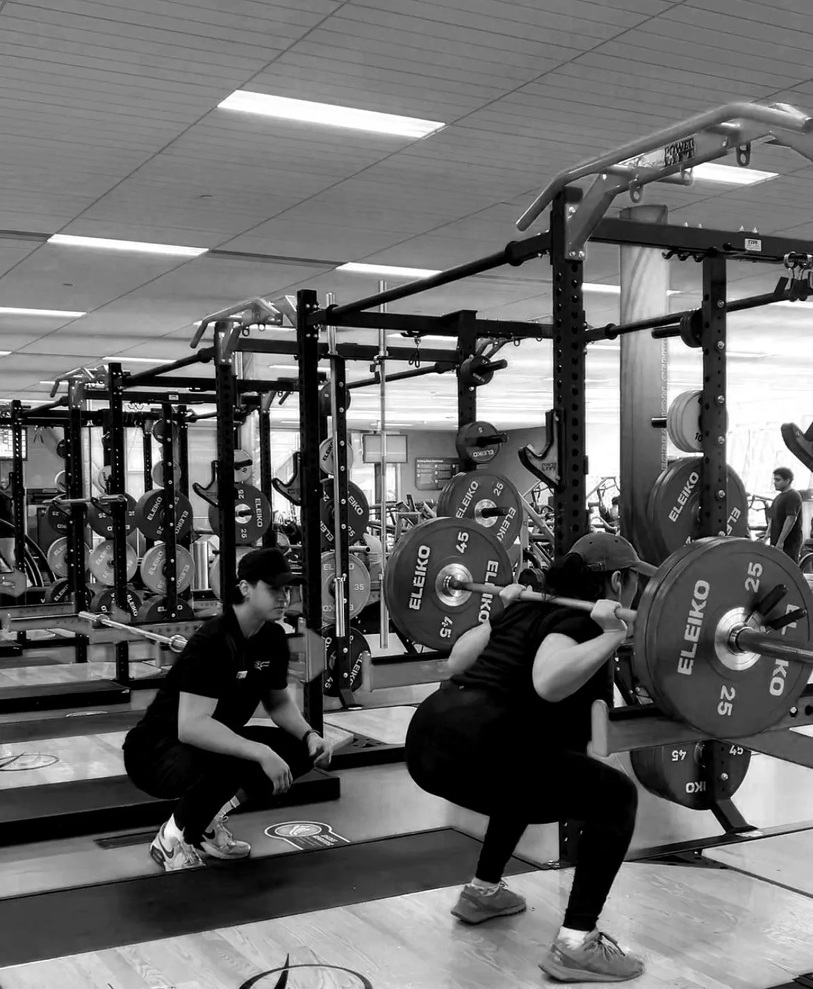 A person performing a squat with a barbell loaded with weights at a gym, with a trainer or staff member nearby and gym equipment in the background.