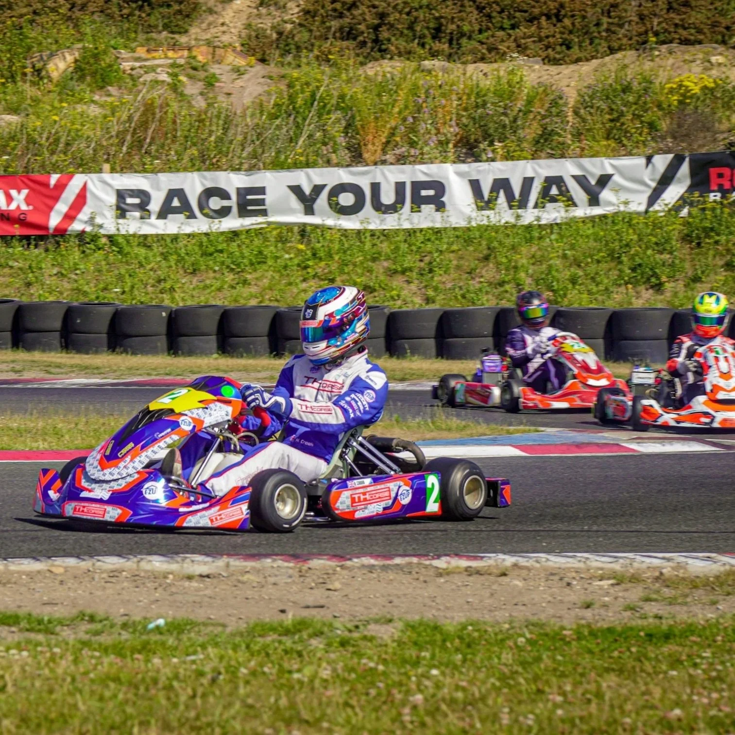 Go-kart racers on a track with tire barriers and a banner that reads 'Race Your Way' in the background.