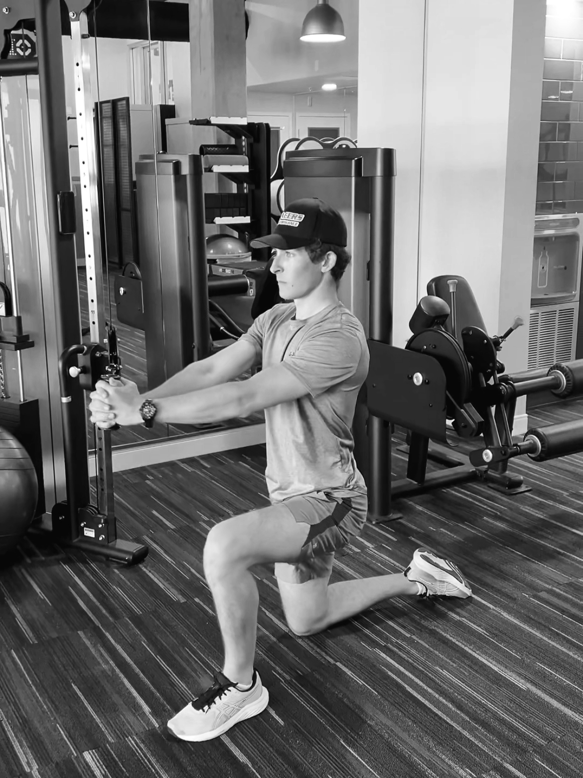 A man wearing a cap, t-shirt, shorts, and sneakers is doing a kneeling exercise with a resistance band at a gym. He is holding onto the band with both hands and pulling it forward while kneeling on one knee. The gym equipment and mirrors are visible in the background.