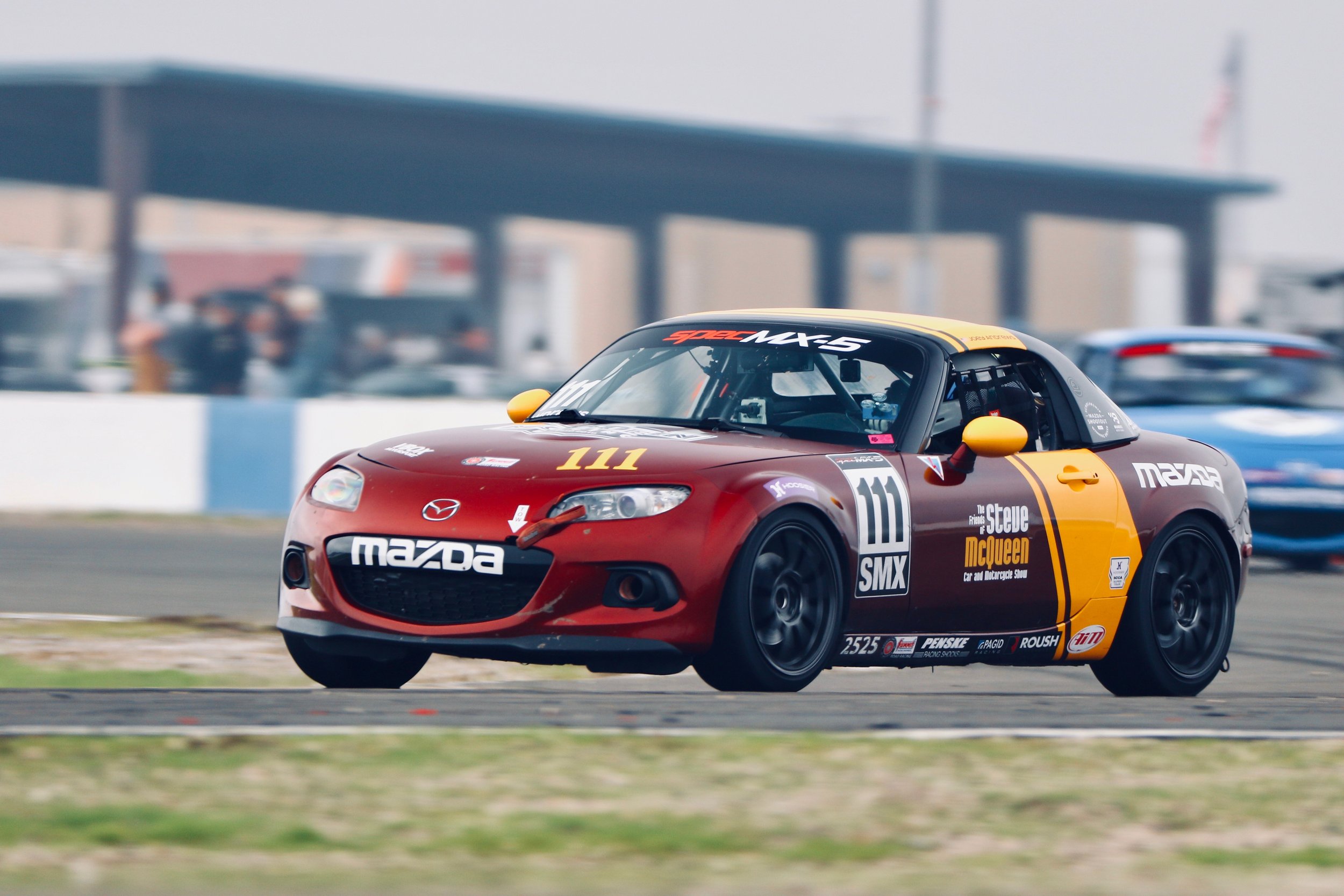 A red Mazda racing car with black and yellow accents speeding on a race track, with racing sponsors and decals on its body and the number 111 displayed on the hood, with a blurred background of other cars and a covered pit area.