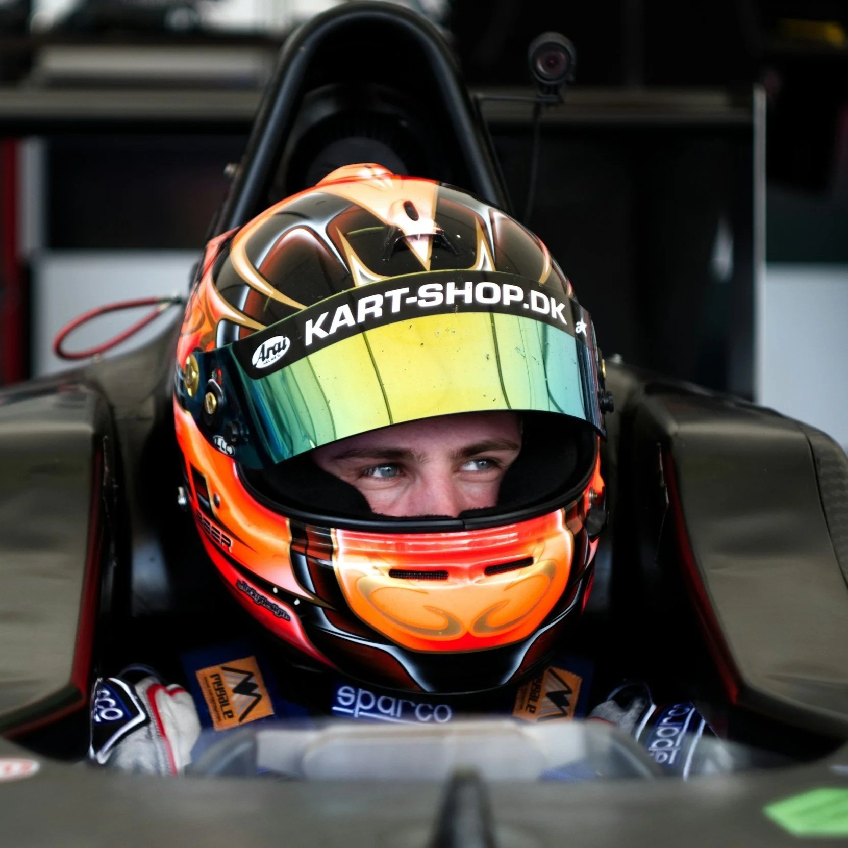 Close-up of a race car driver wearing a helmet with a reflective visor, sitting in a race car with a full cockpit. The helmet has black, orange, and gold colors, and the visor reflects the surroundings. The driver appears focused. The helmet's brim features a
