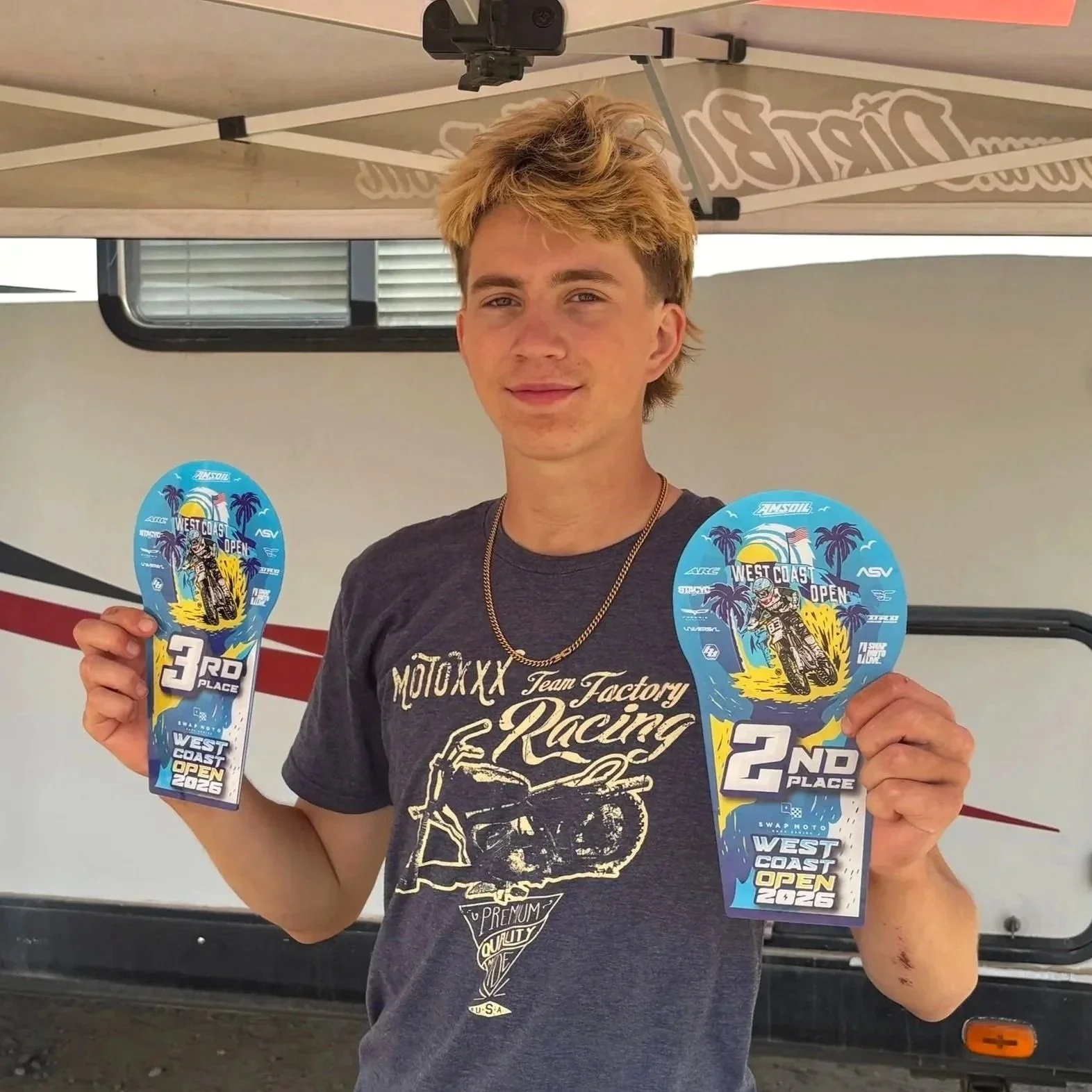 Young man with blond hair smiling, holding two colorful awards for third and second place, standing in front of a vehicle, at the West Coast Open 2026 motocross event, under a canopy.