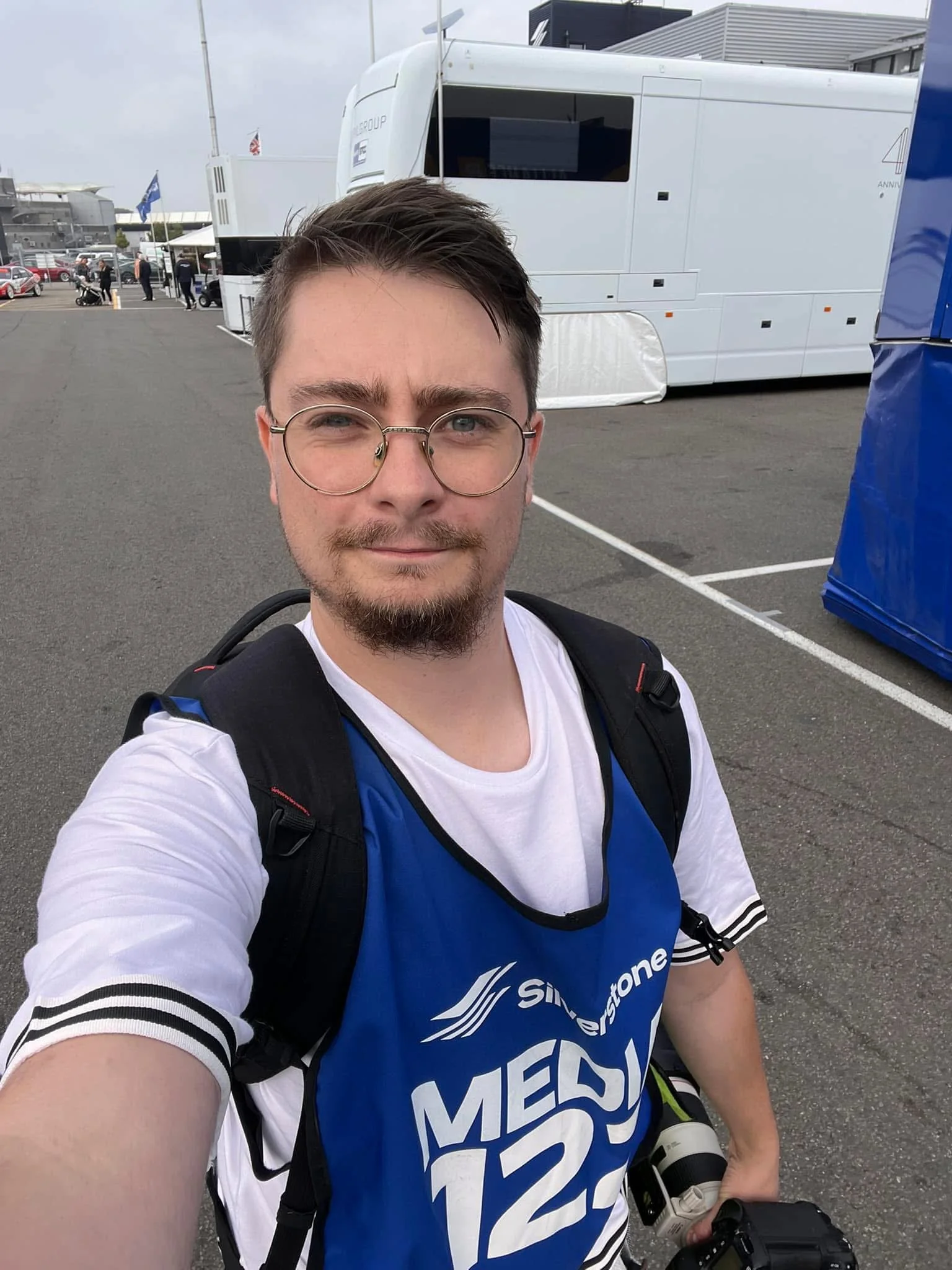 A young man with glasses and a backpack taking a selfie in a parking lot, with a large white mobile broadcasting truck in the background, several people, and parked cars.
