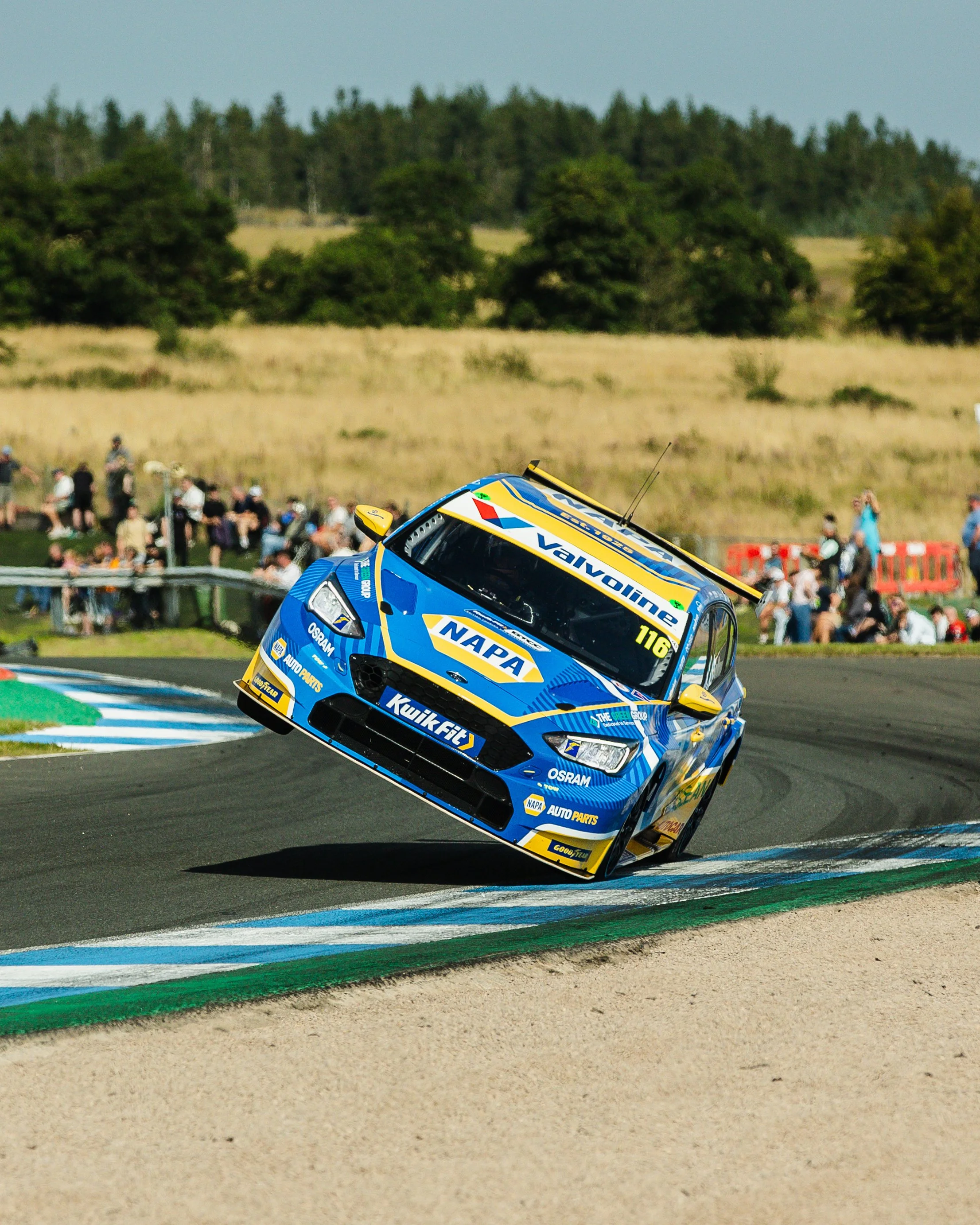 A race car with sponsorship stickers is airborne, tilted at an angle, on a race track with lap markings and a crowd of spectators in the background.
