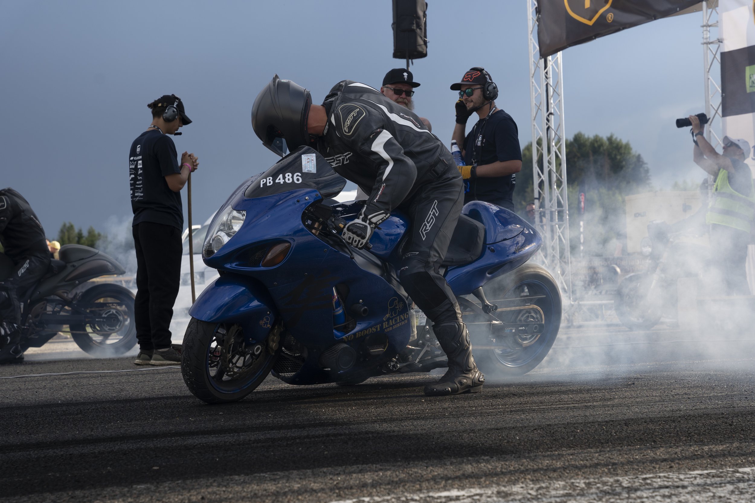 Motorcycle rider in racing suit and helmet performing burnout on a blue sportbike at a racetrack, with crew members and officials nearby, smoke and a cloudy sky in the background.