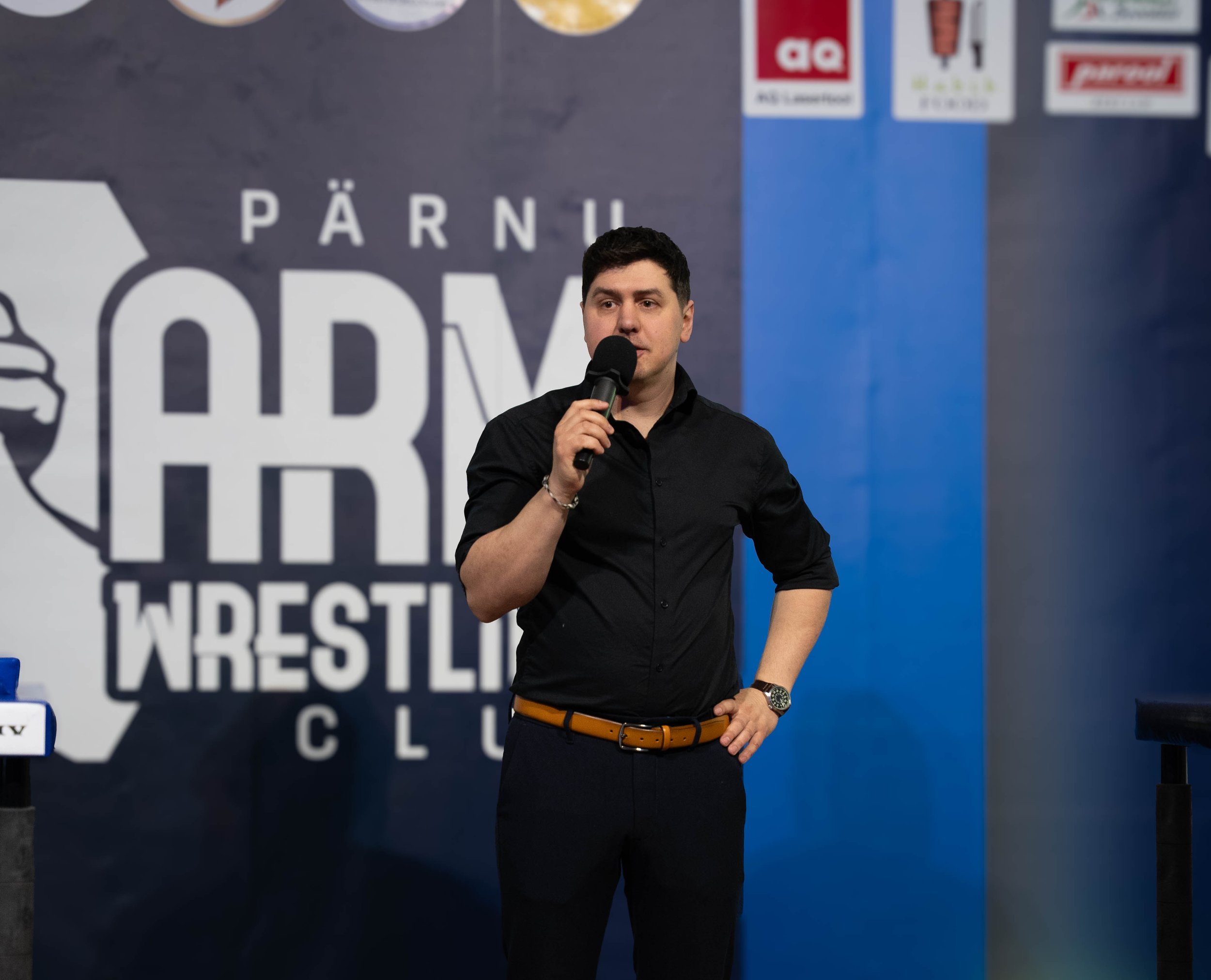 A man in black shirt holding a microphone speaking at a wrestling event, with a backdrop displaying the words "Pärnu Wrestling Club."
