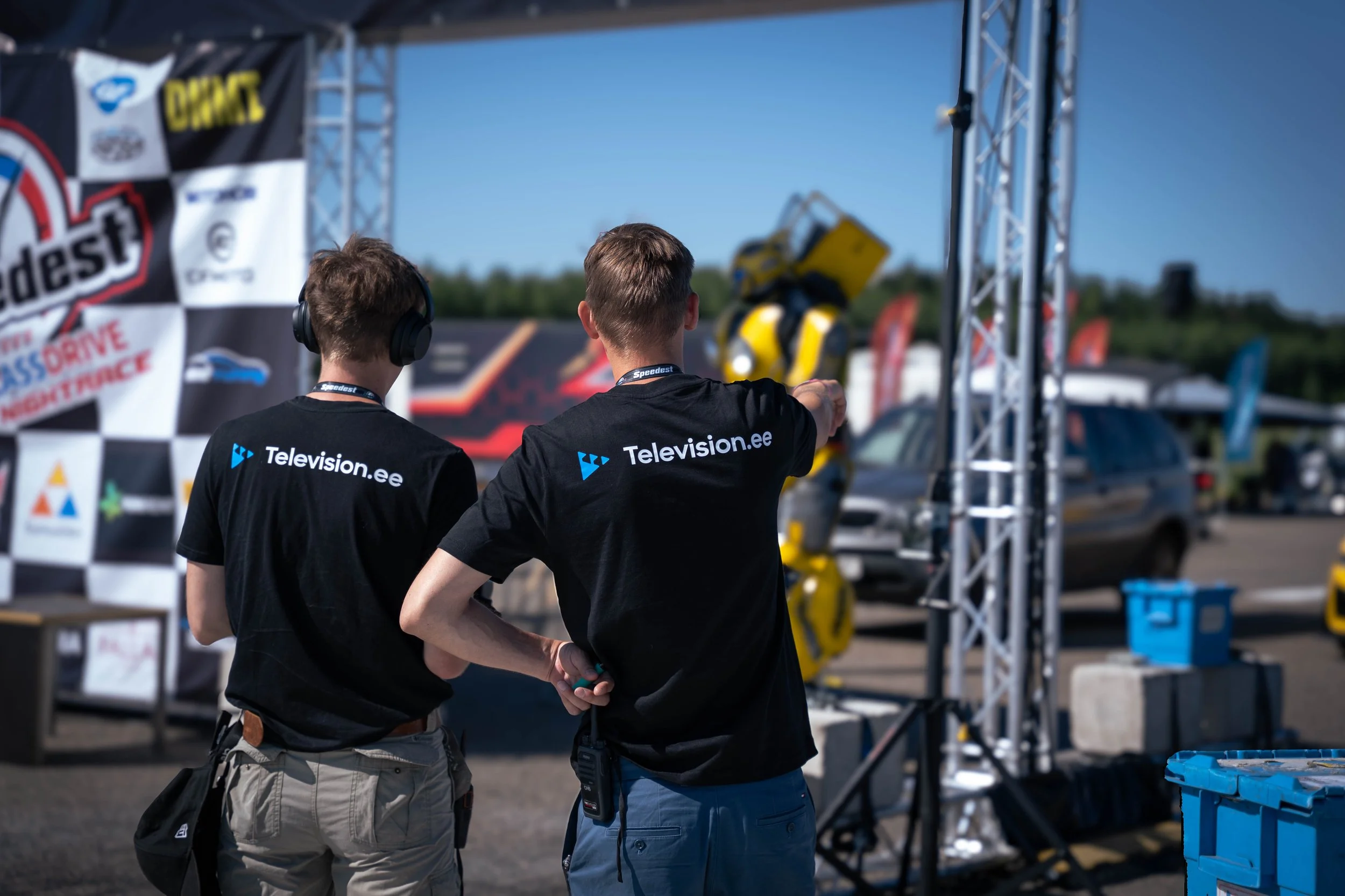 Two men wearing black T-shirts with 'Television.ee' printed on the back are standing outdoors next to a metal frame with equipment, looking towards a yellow and black robot.