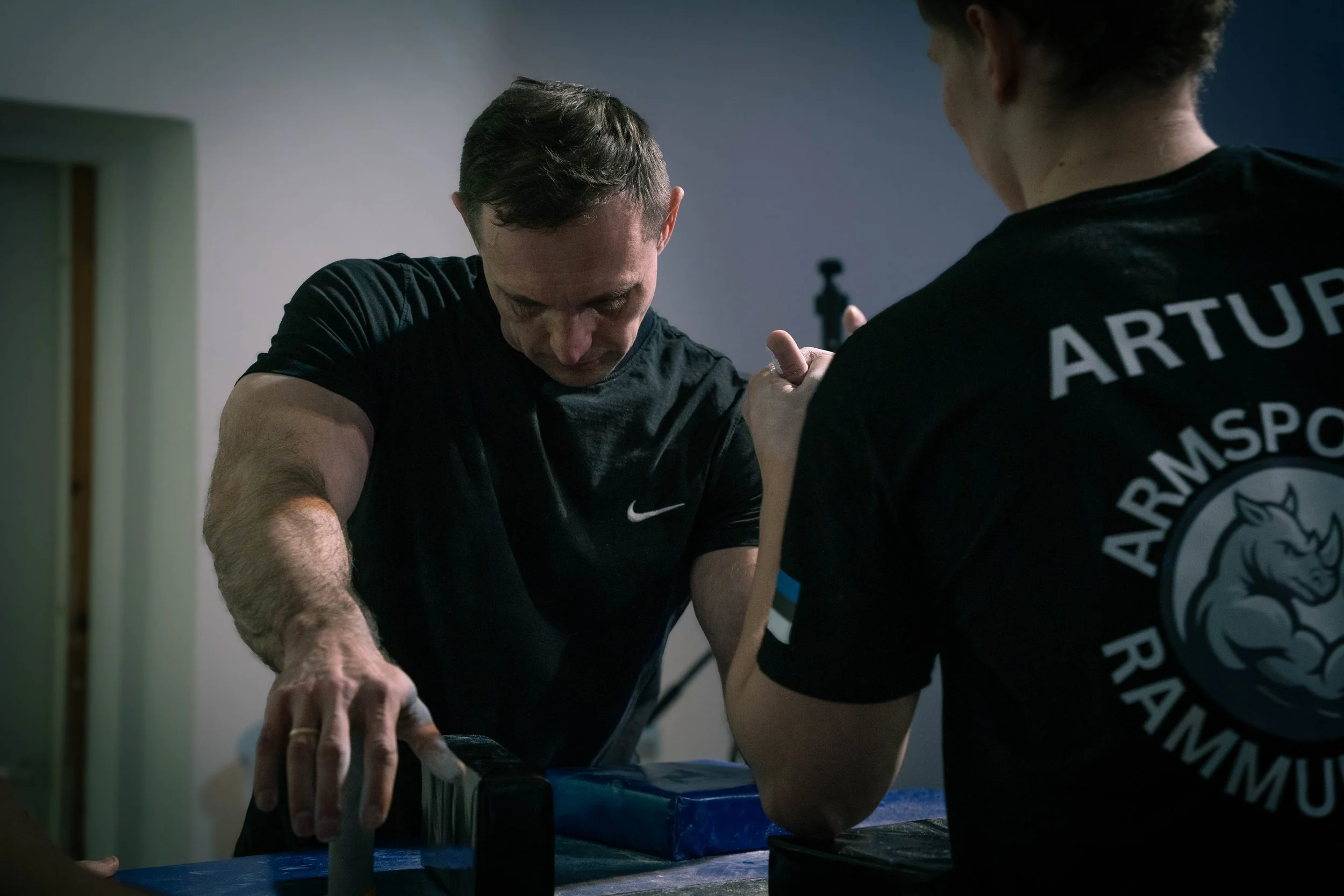 Two men arm wrestling indoors, one with a Nike shirt and the other with a shirt that reads 'ARTUR ARMSPORT RAINMU'.